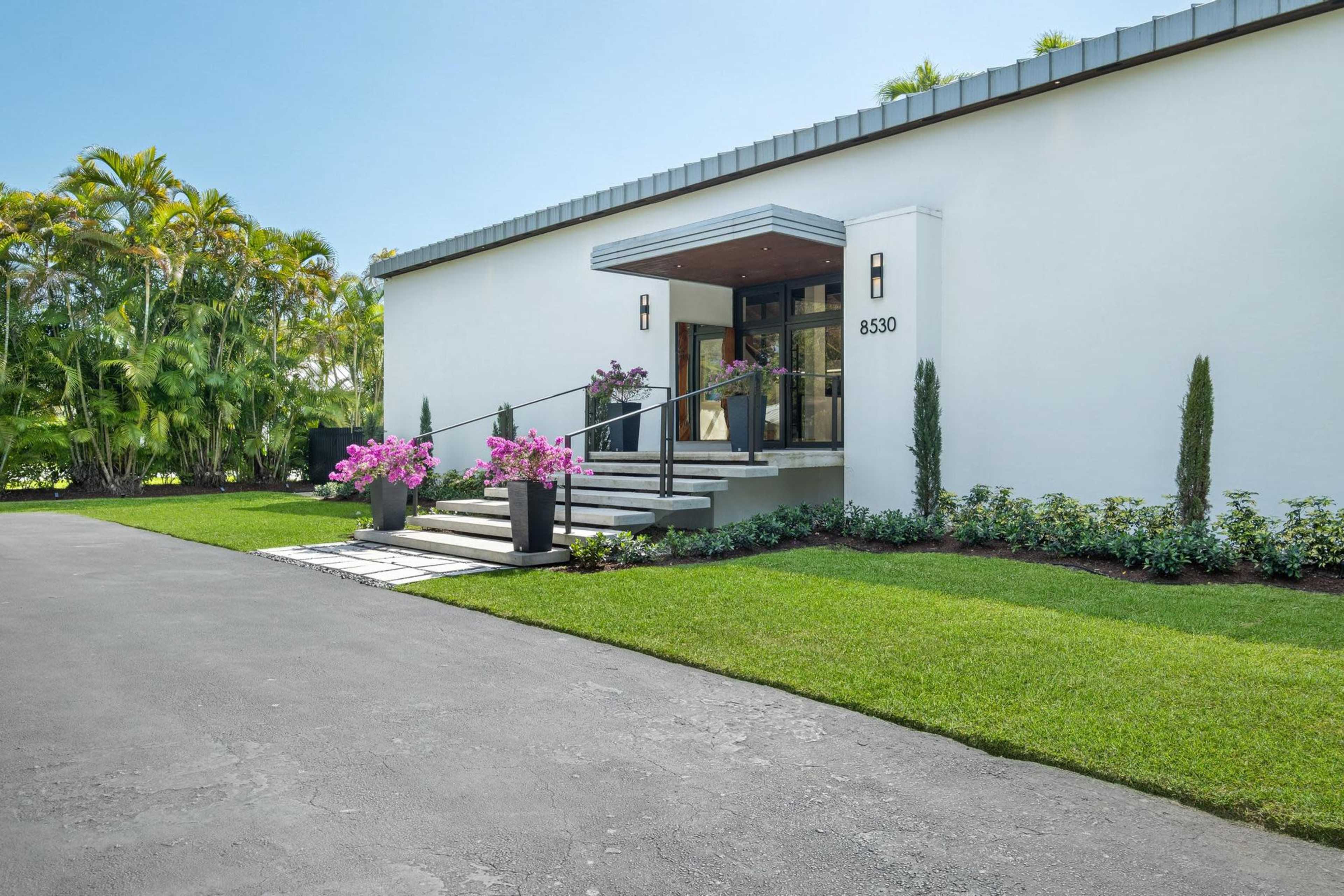The image shows a modern white house with a landscaped entrance, featuring a stone path, planters with pink flowers, and a prominent front door.