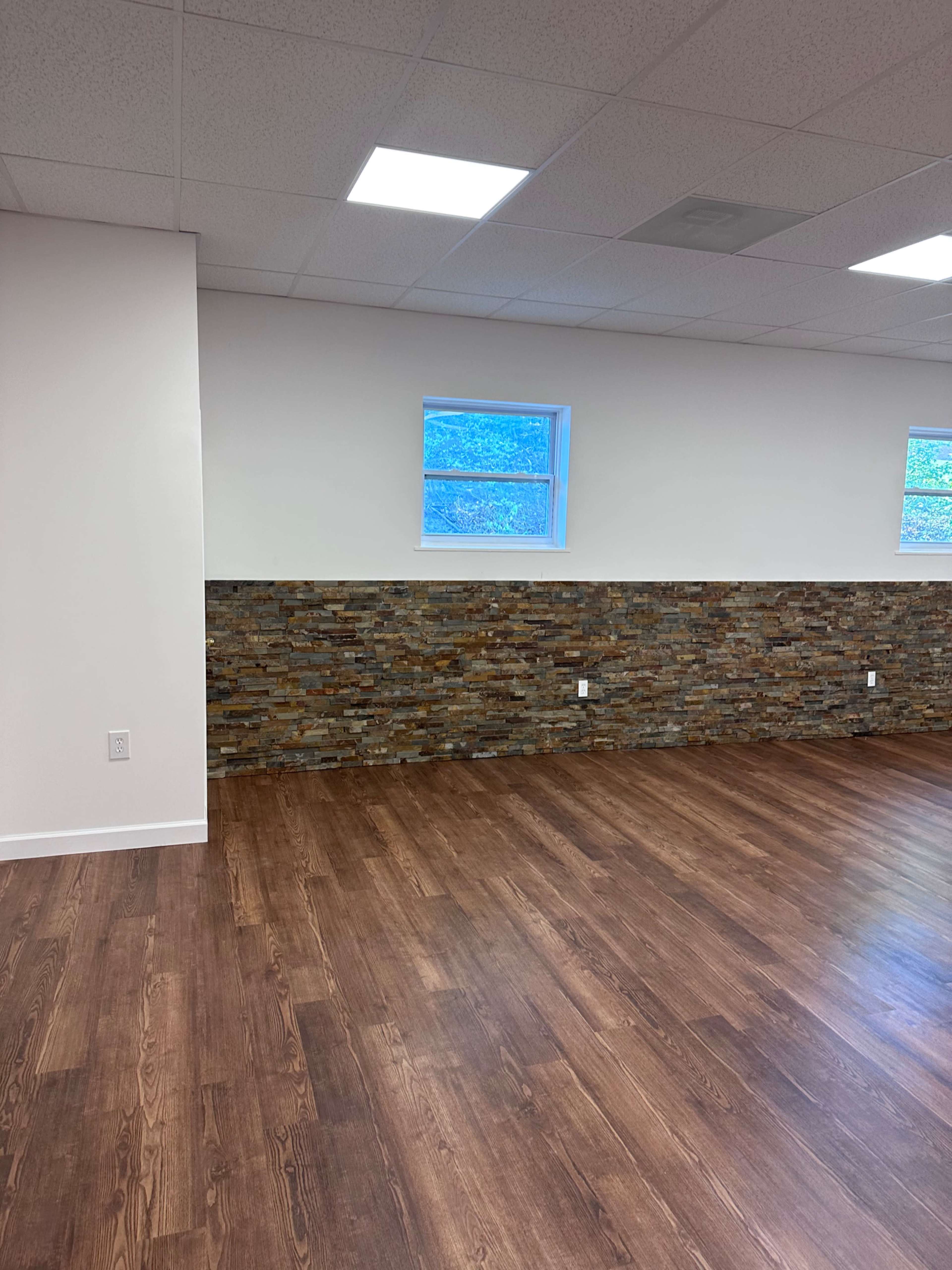 The image shows a spacious room featuring a wooden floor, a stone accent wall, and a few small windows allowing natural light to enter.