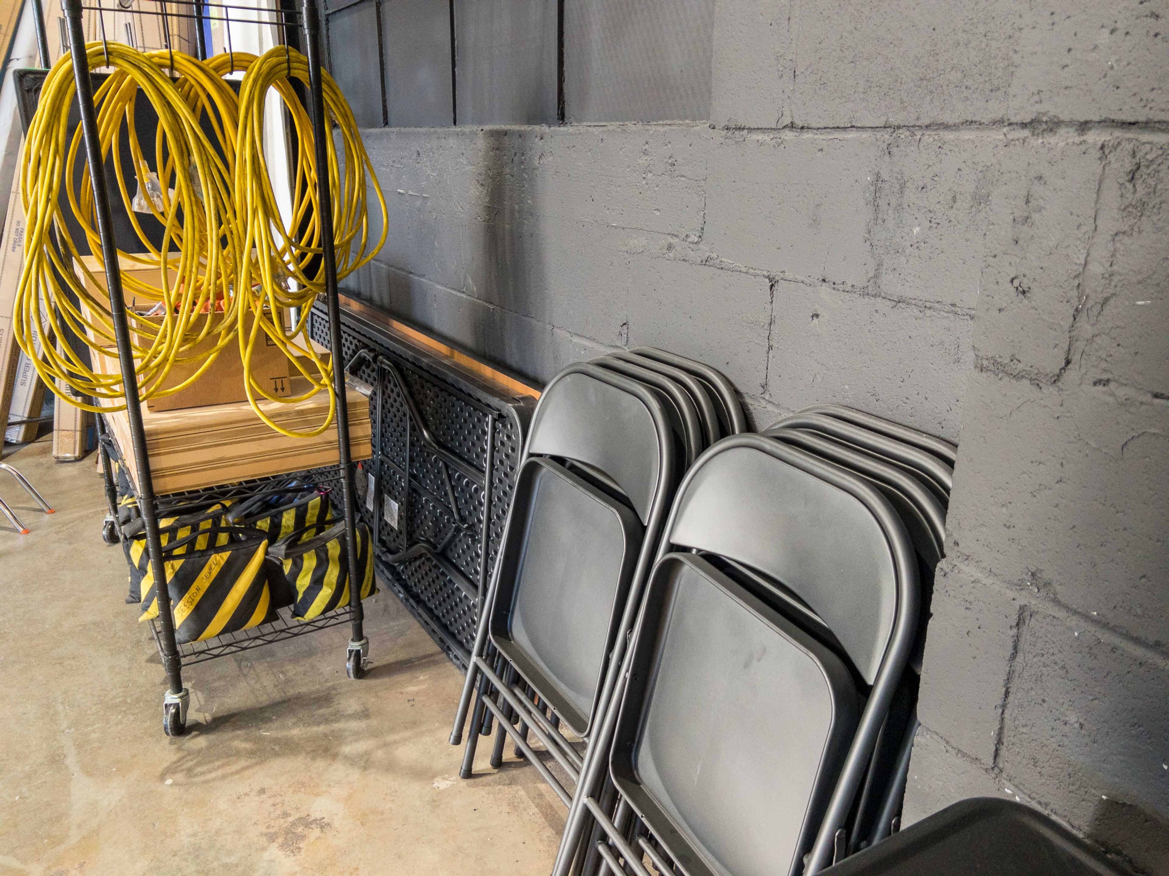 A stack of black folding chairs is stored against a gray wall next to a cart with yellow extension cords and wooden planks.