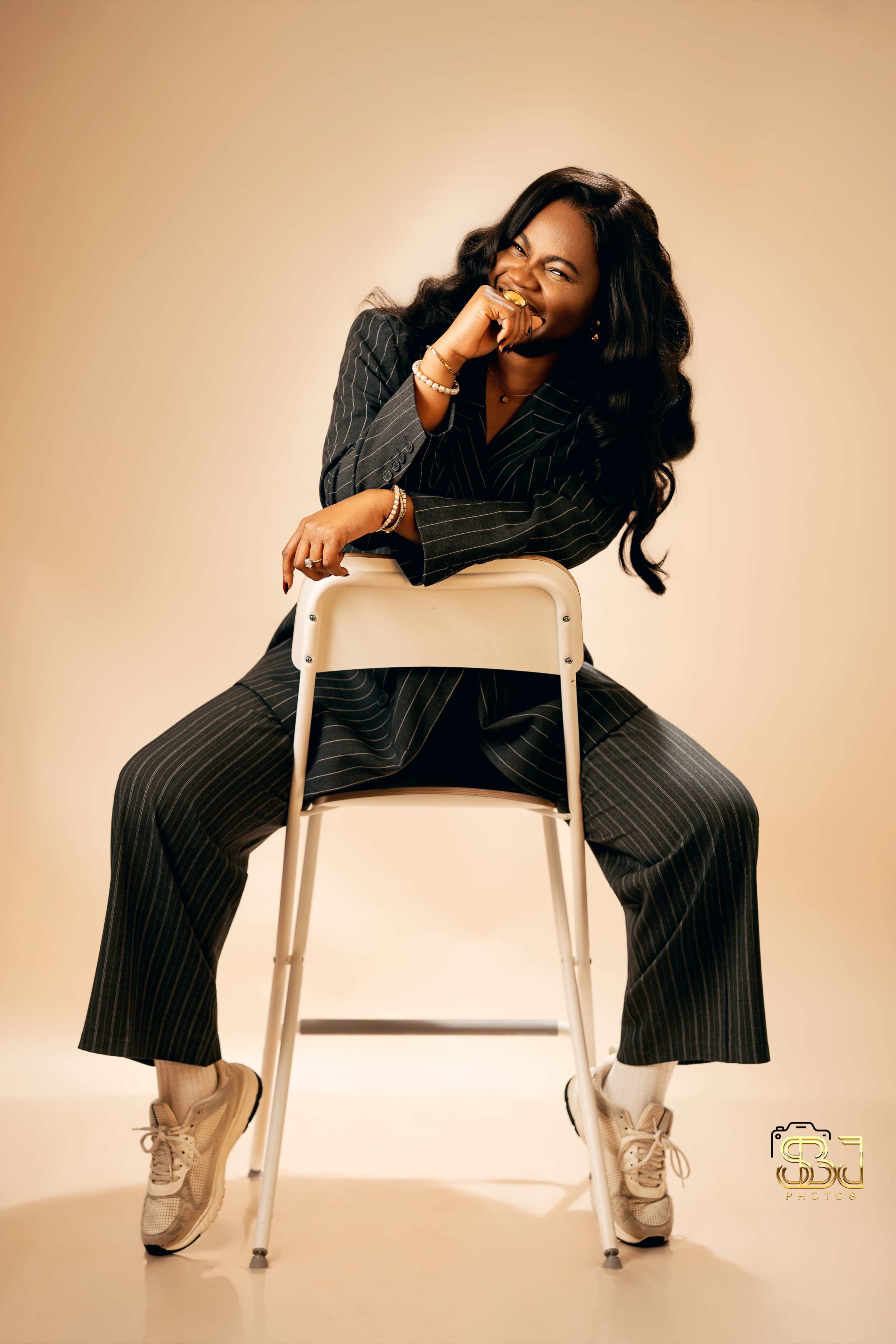 A person in a pinstripe suit is sitting on a chair, laughing while holding their hand to their mouth.