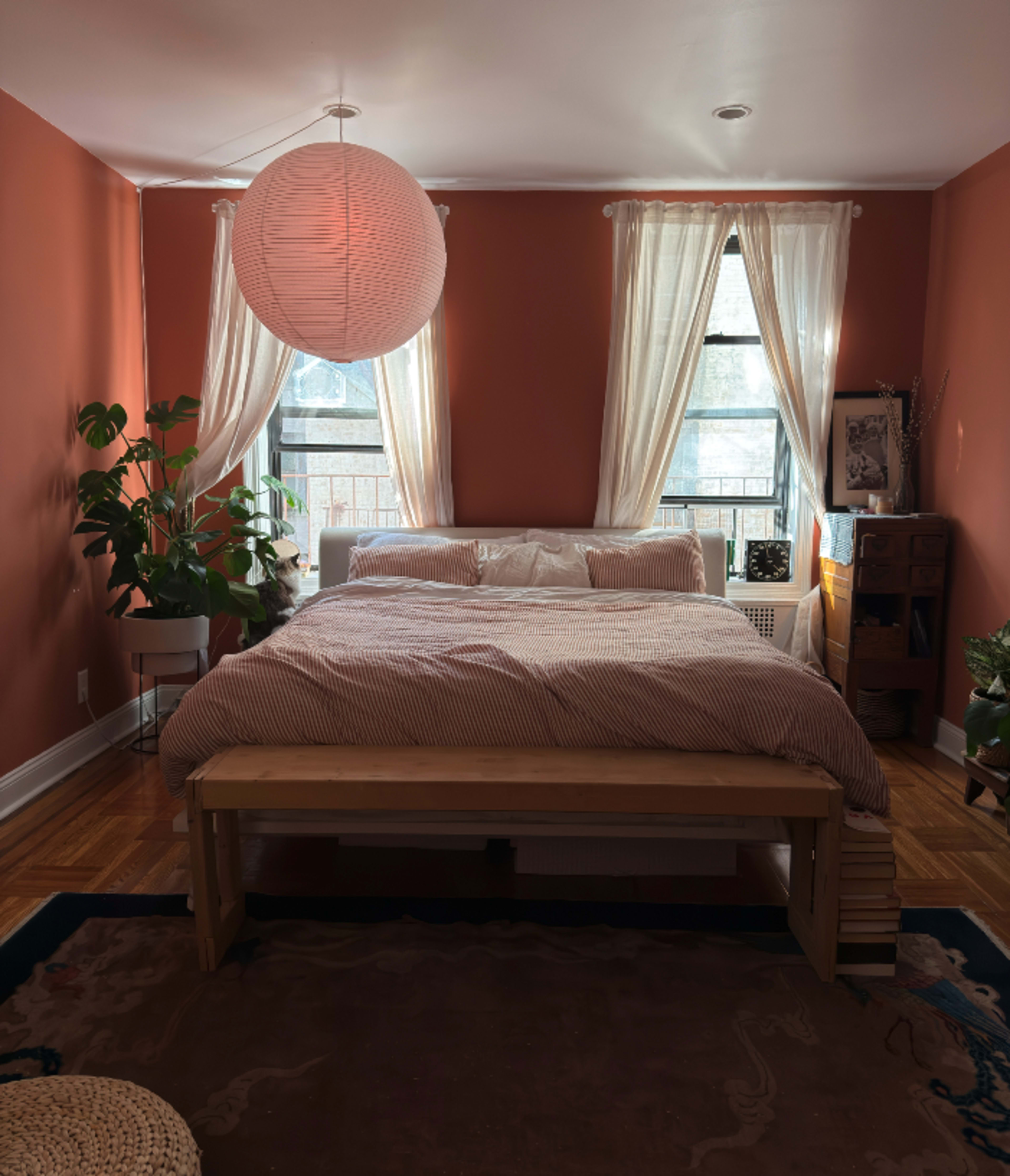 The image shows a cozy bedroom with a bed, large windows covered by sheer curtains, and a pendant light hanging from the ceiling, complemented by plants and wooden furniture.