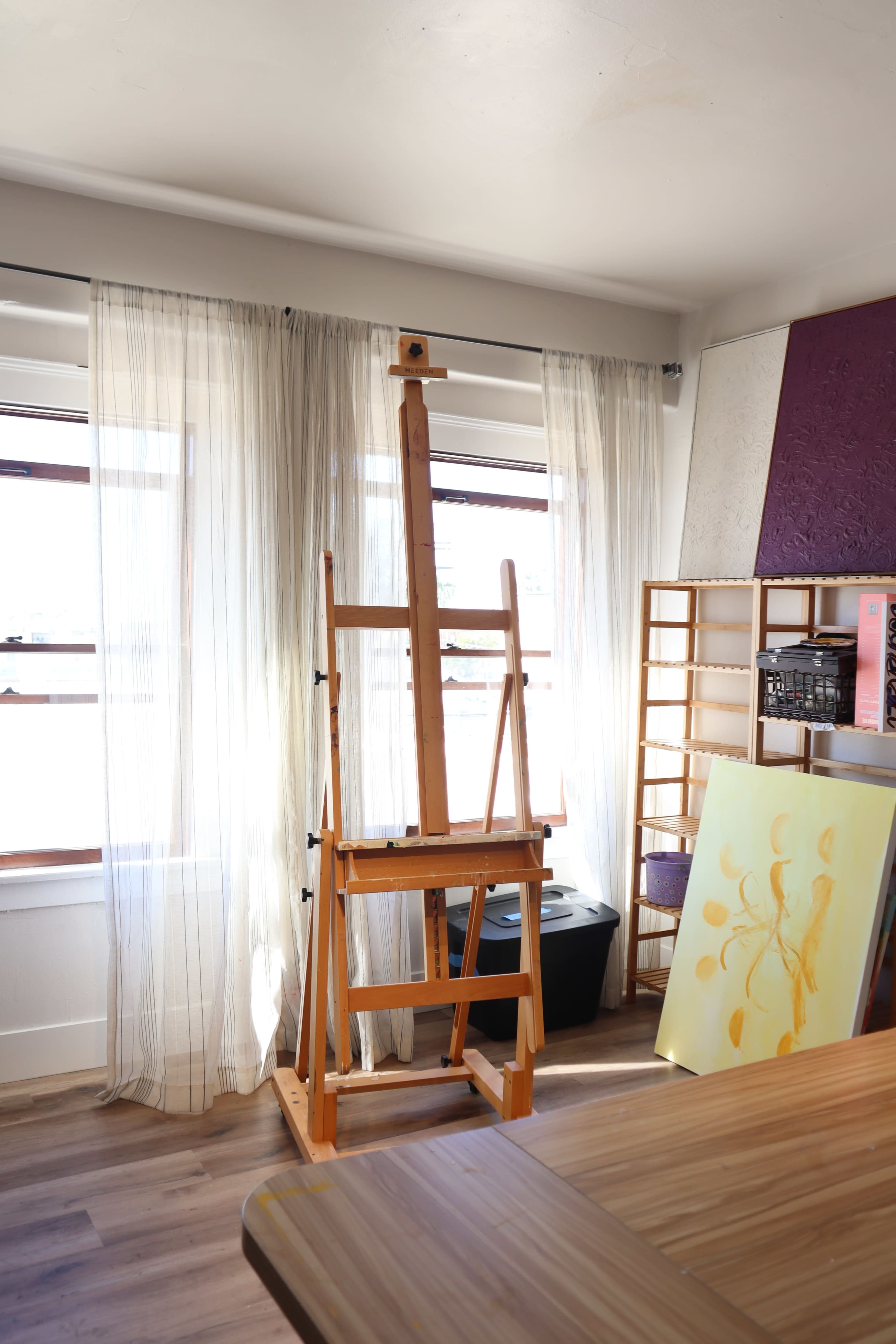 A wooden easel stands beside large windows, with sheer curtains and an artist's palette leaned against a nearby wall.