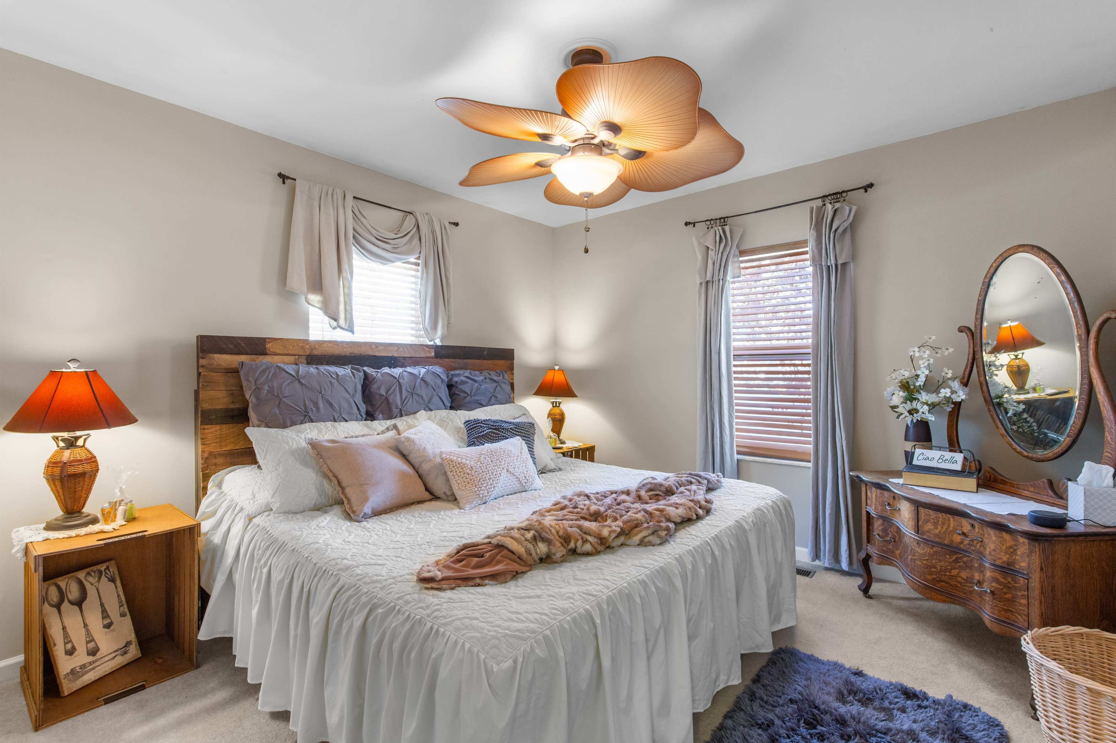 The image shows a cozy bedroom with a large bed, a decorative ceiling fan, bedside lamps, and a wooden dresser with a round mirror.