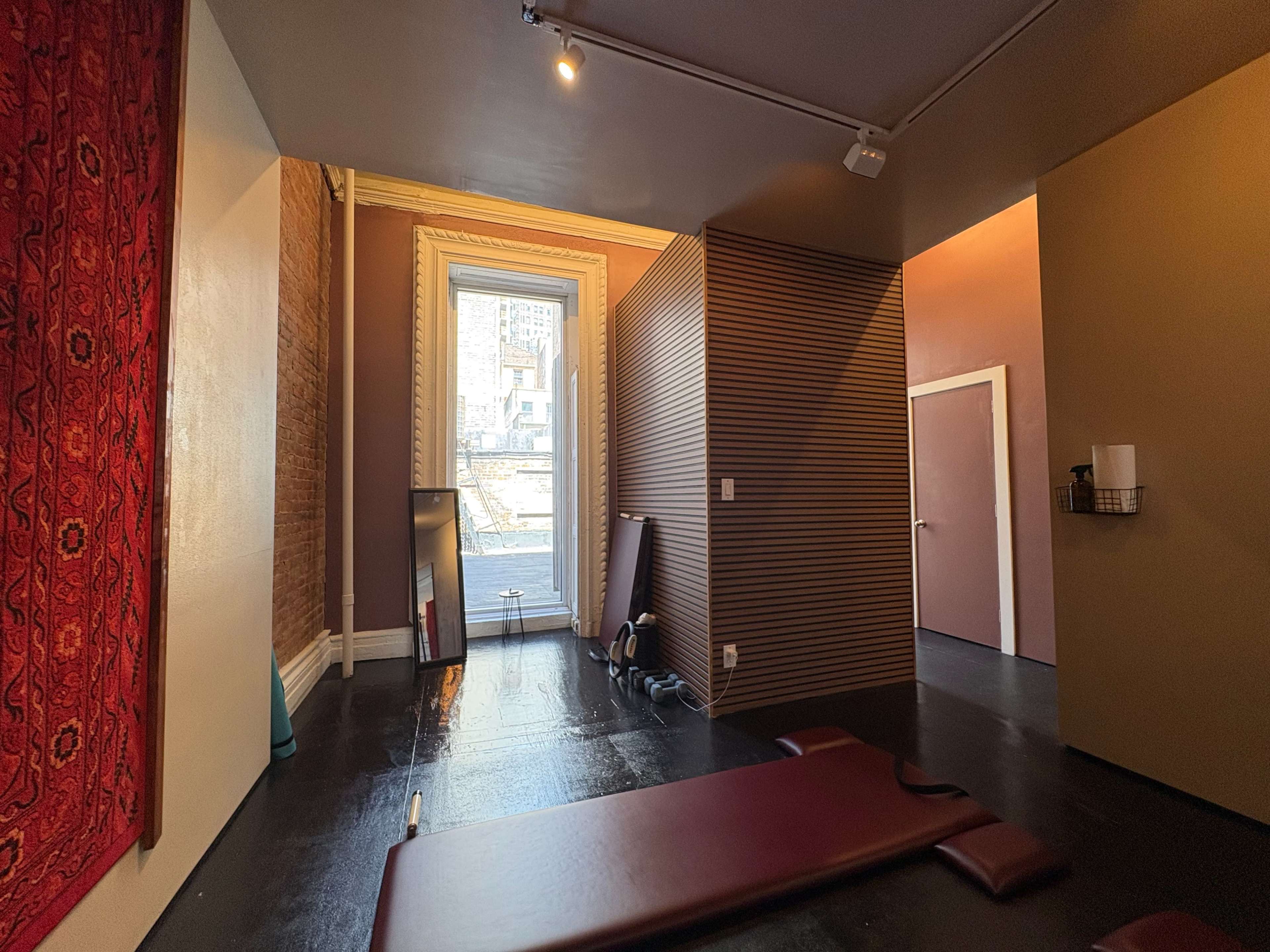 The image shows a spacious interior with wooden floors, a red decorative wall hanging, and a corridor leading to a window.