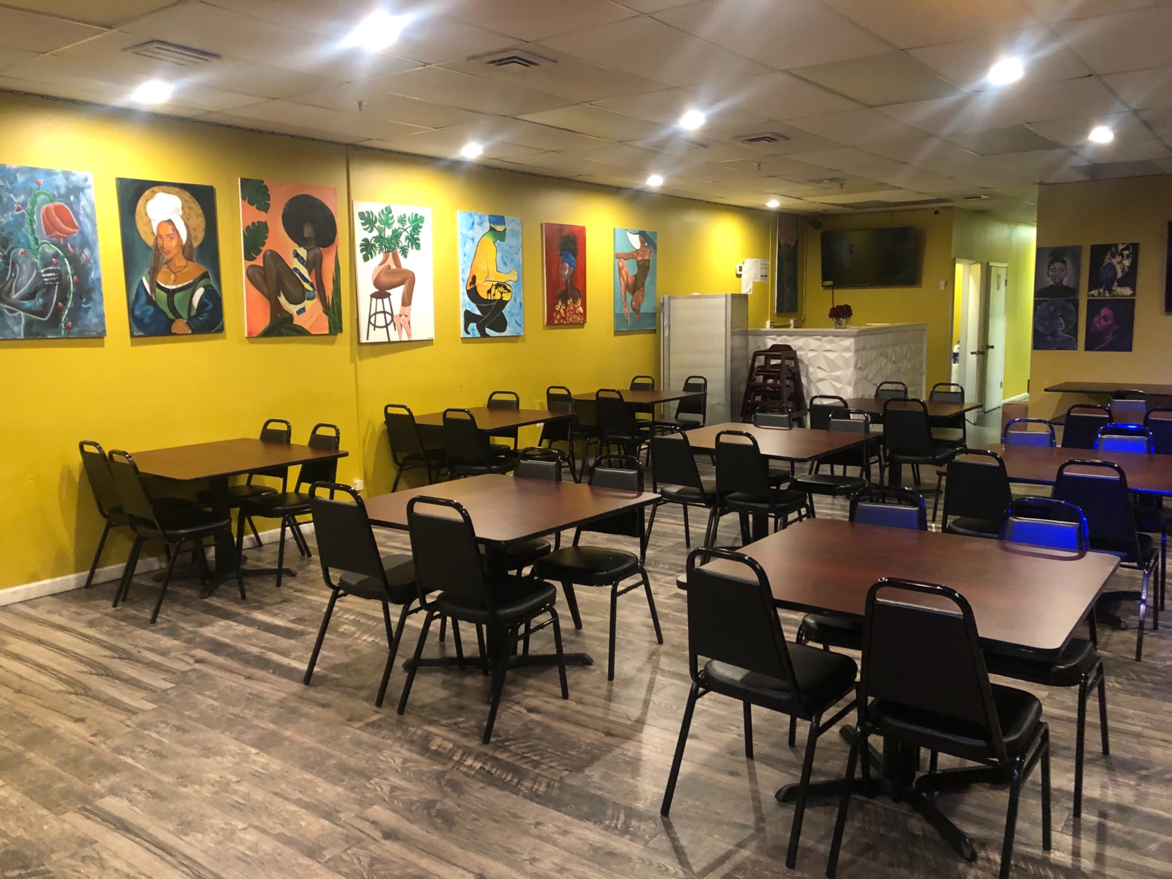 A brightly colored dining area with multiple empty tables and chairs arranged neatly, adorned with various paintings on the yellow walls.
