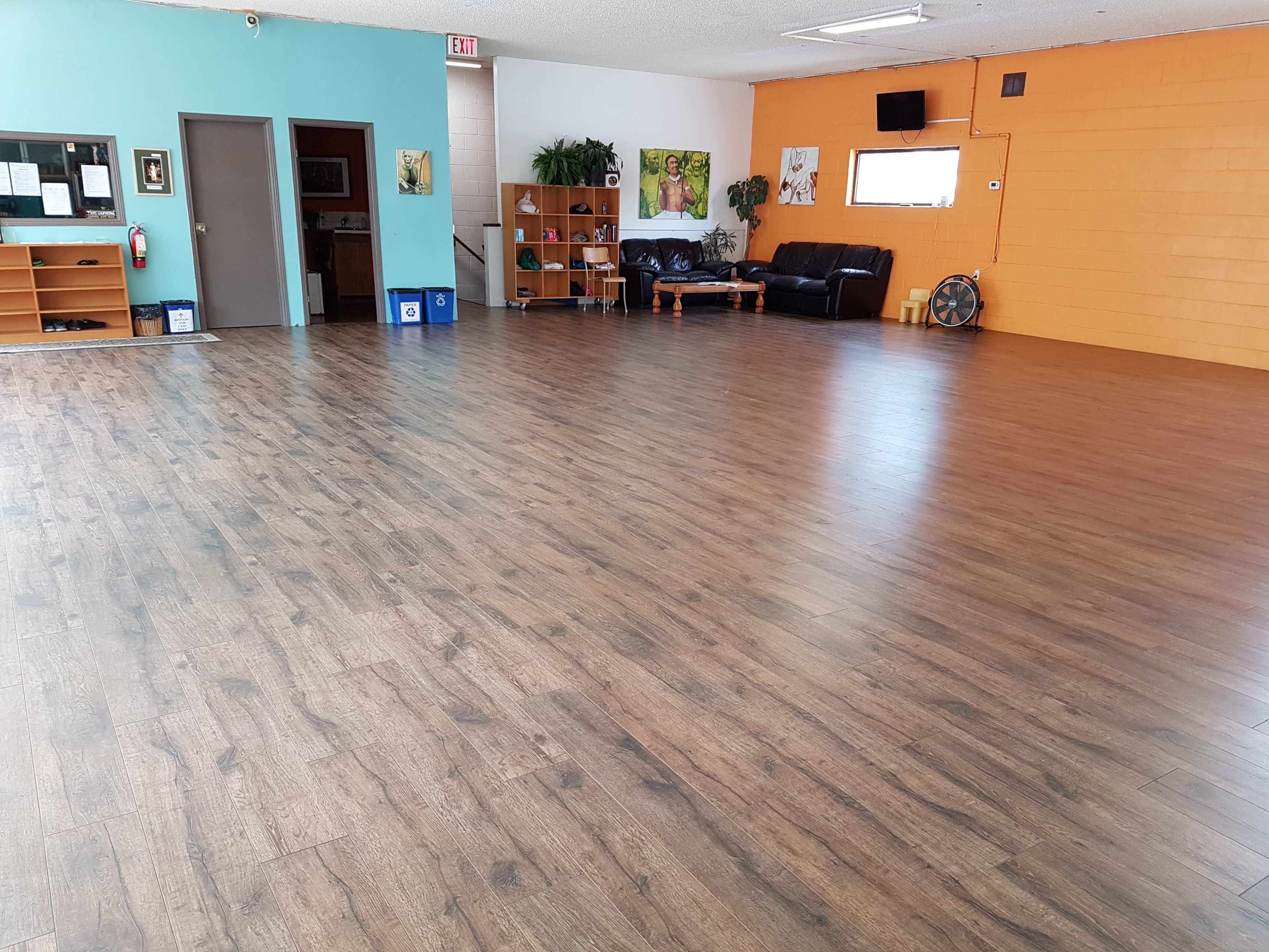 A spacious interior room with wooden flooring, featuring a light blue wall on one side and an orange wall on the other, along with some furniture and plants.