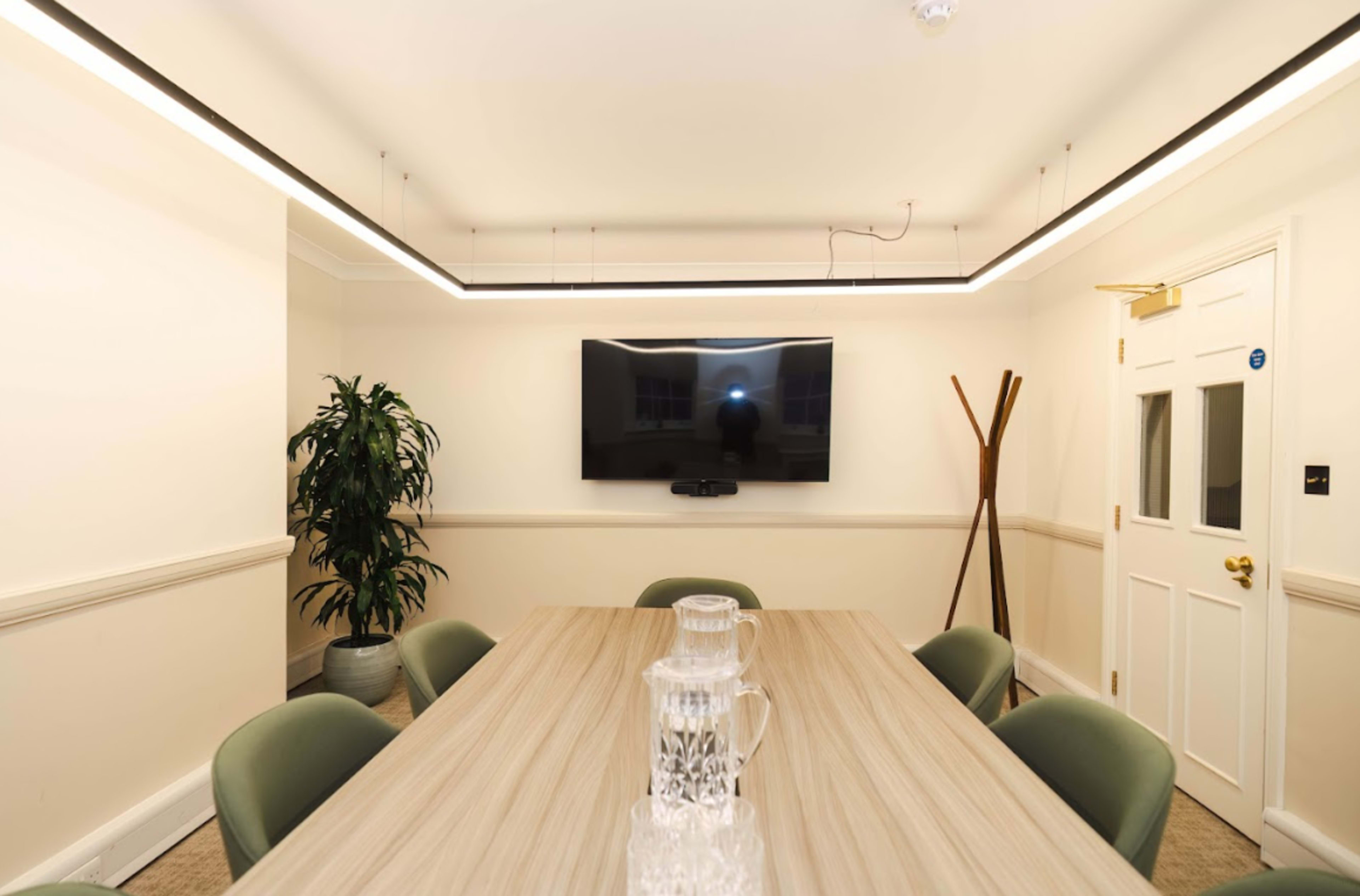 A modern conference room features a long wooden table surrounded by green chairs, with a television mounted on the wall and a plant in the corner.