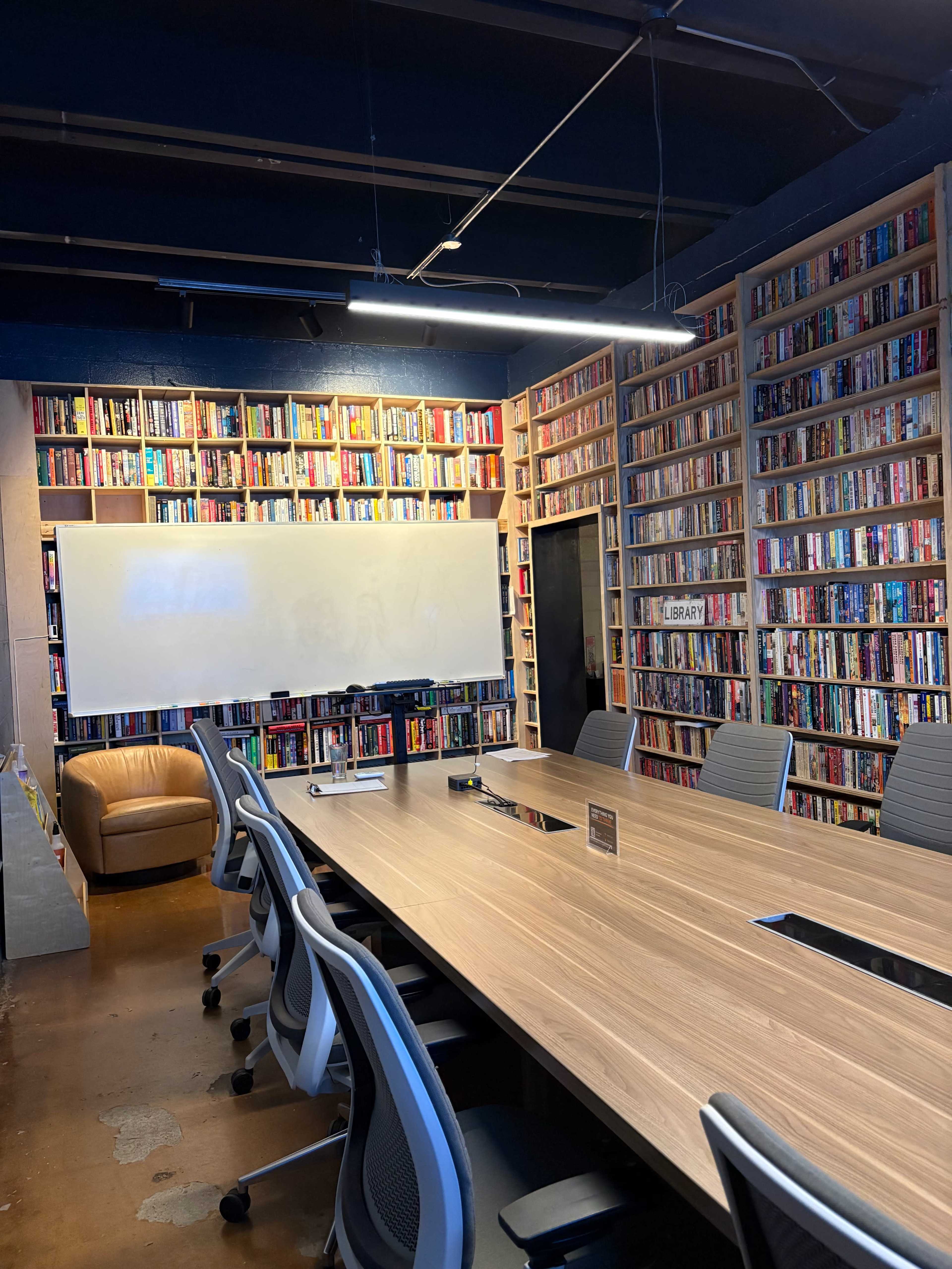 The image shows a meeting room with a long wooden table surrounded by office chairs, a whiteboard, and bookshelves filled with various books lining the walls.