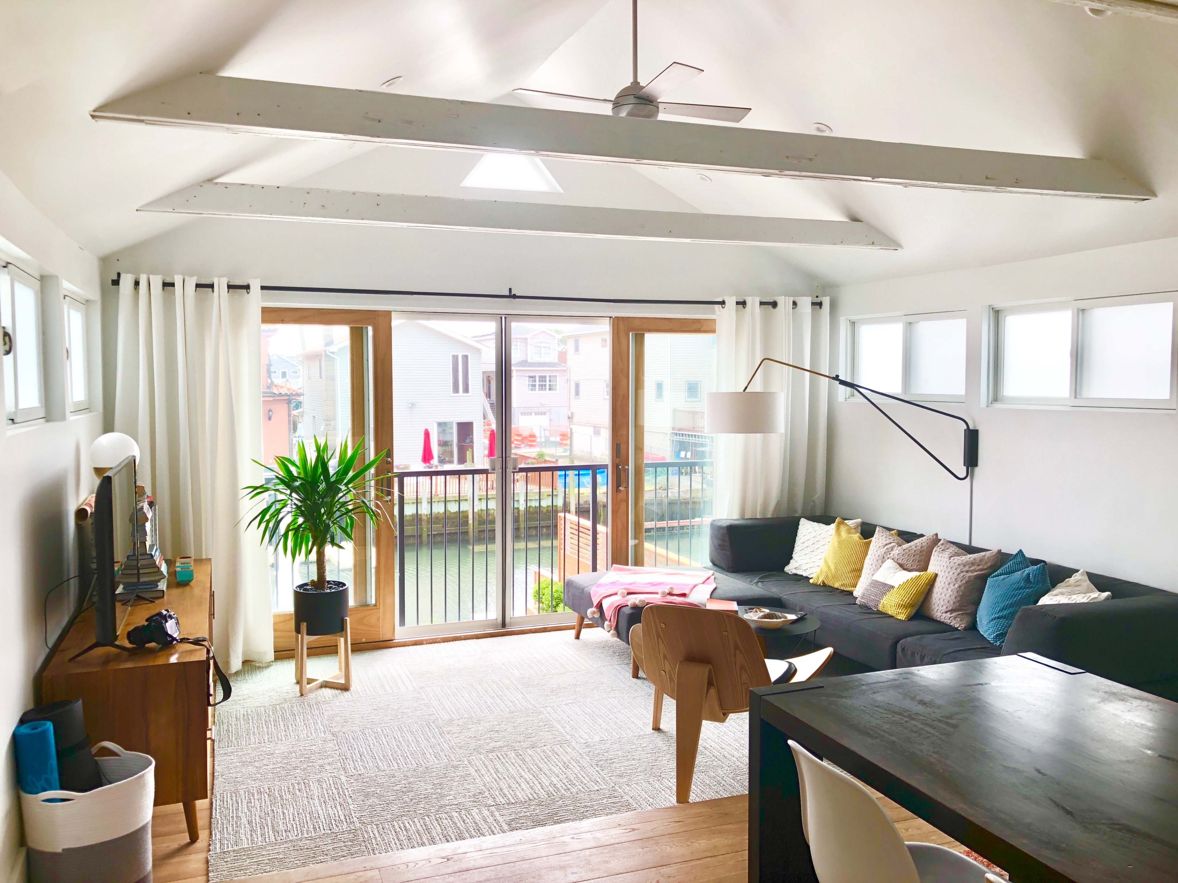 The living room features a sectional sofa, a dining table, and large windows overlooking a deck with a view of nearby houses.