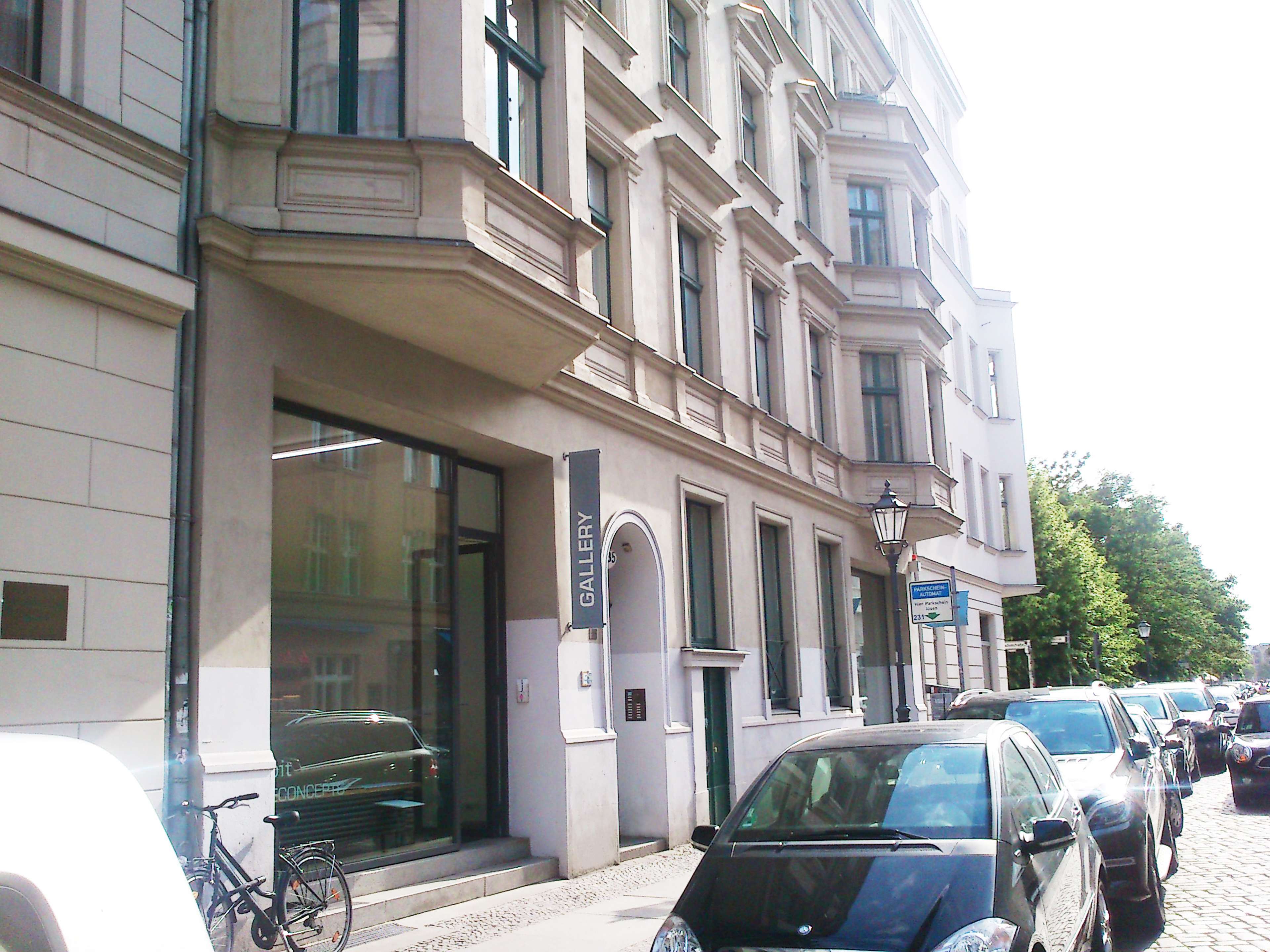The image shows a street view of a multi-story building with a large glass window displaying a sign that reads "GALLERY," surrounded by parked cars and trees.