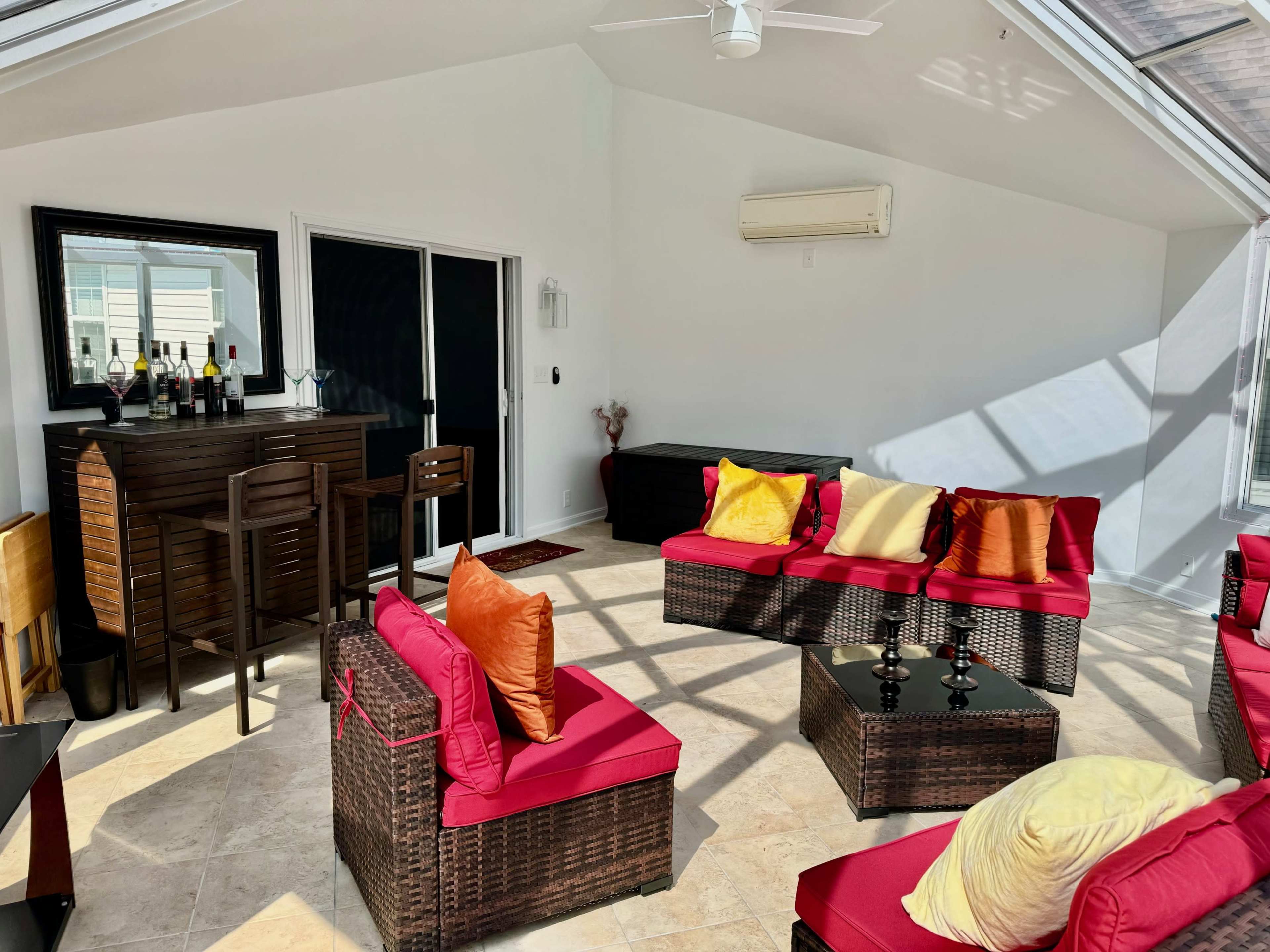 A sunlit room features wicker furniture with red cushions, a bar area with bottles on display, and a large glass door leading outside.