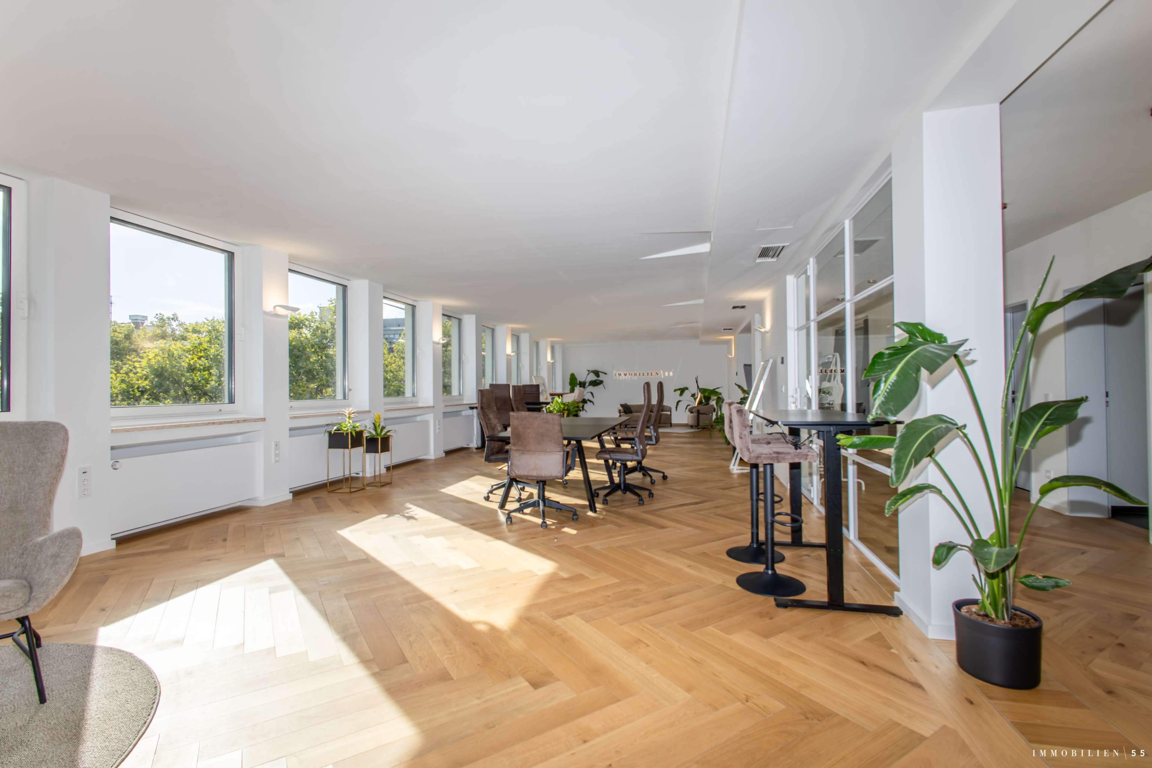 The image shows a bright, modern office space with wooden flooring, large windows, and several seating areas including chairs and small tables.