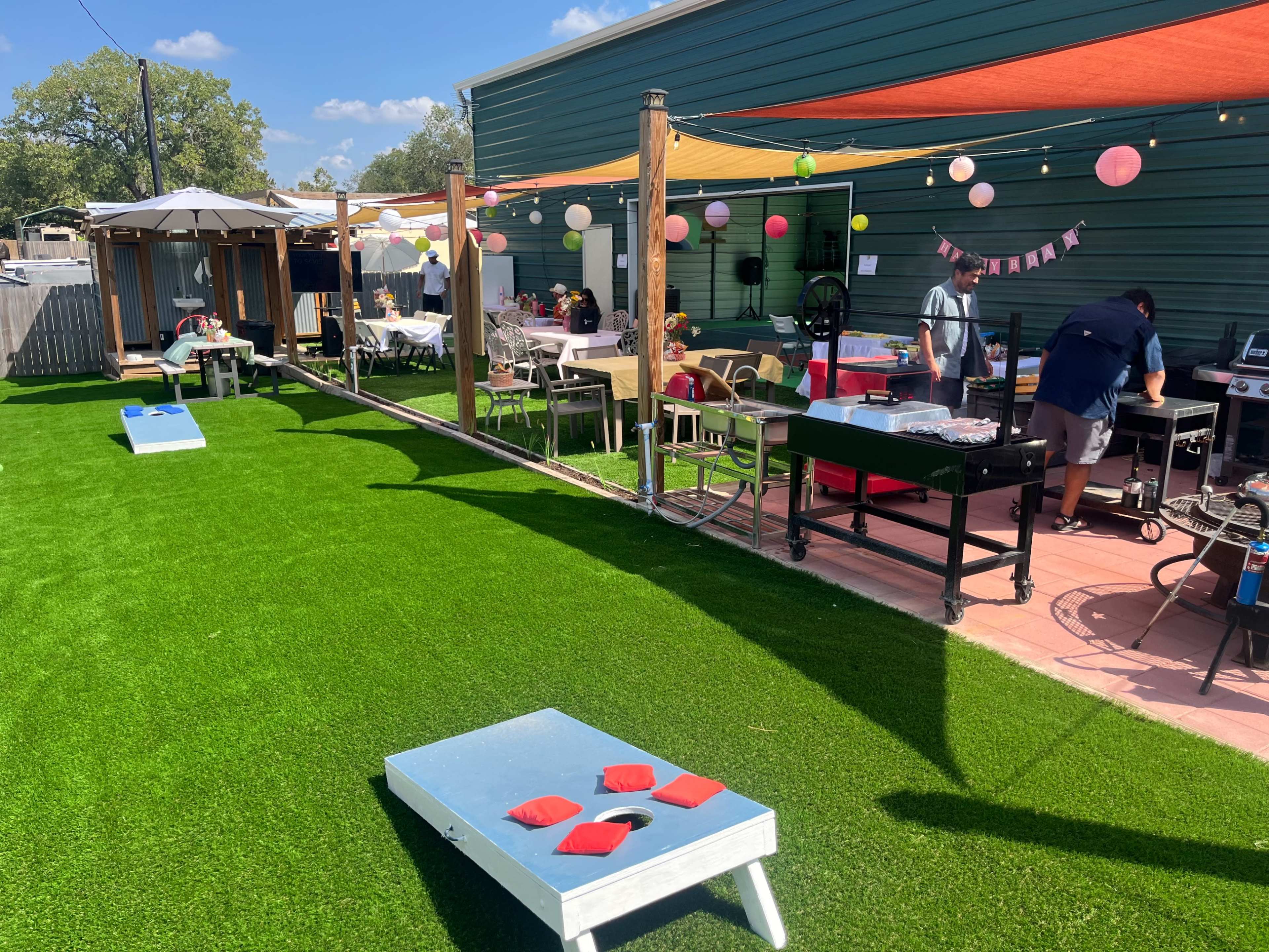 The image shows a backyard gathering with a grassy area featuring cornhole boards, a barbecue setup, dining tables, and colorful decorations.