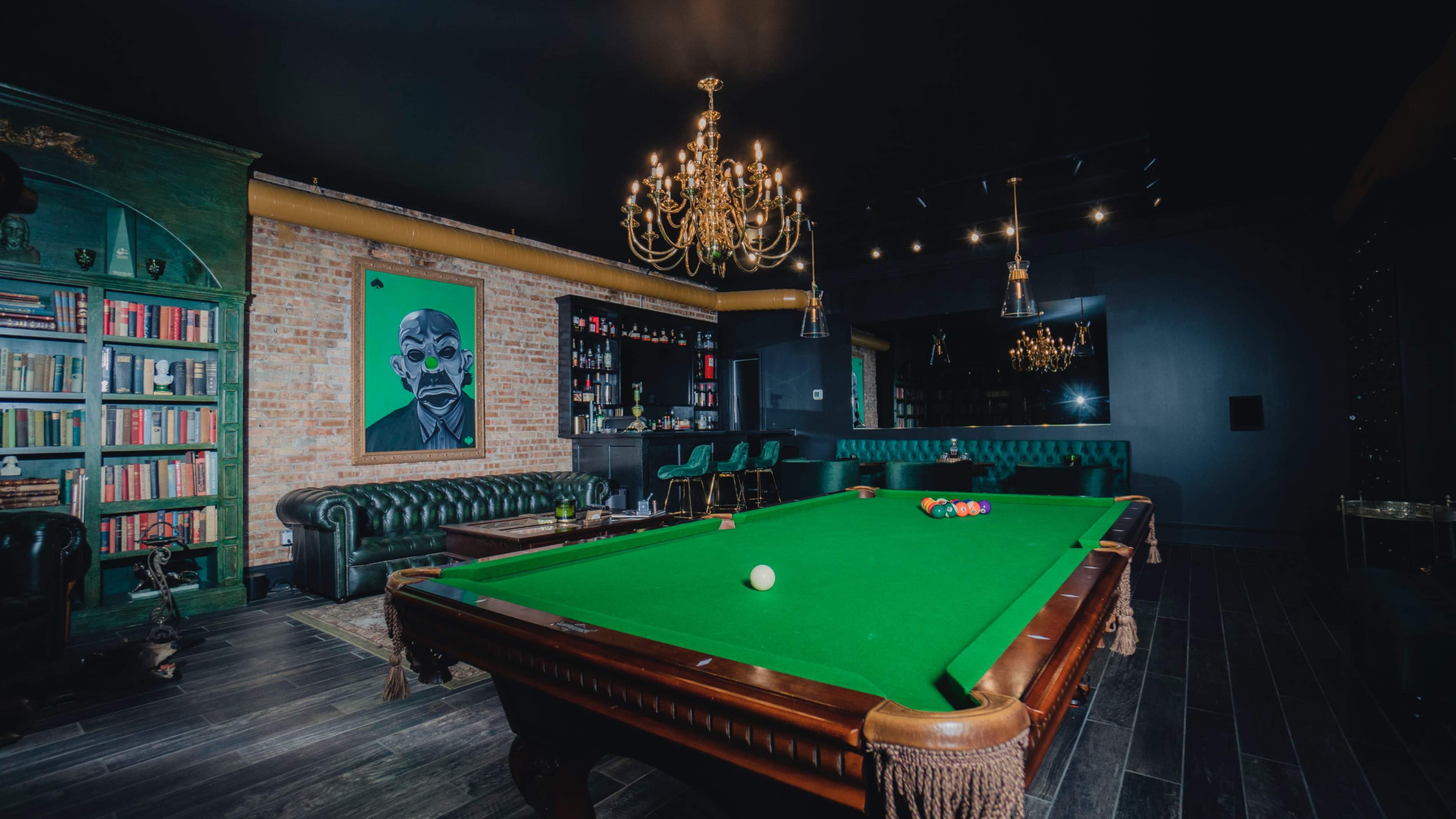 A spacious game room features a green billiards table, a classic leather sofa, shelves lined with books, and a striking portrait on the wall.