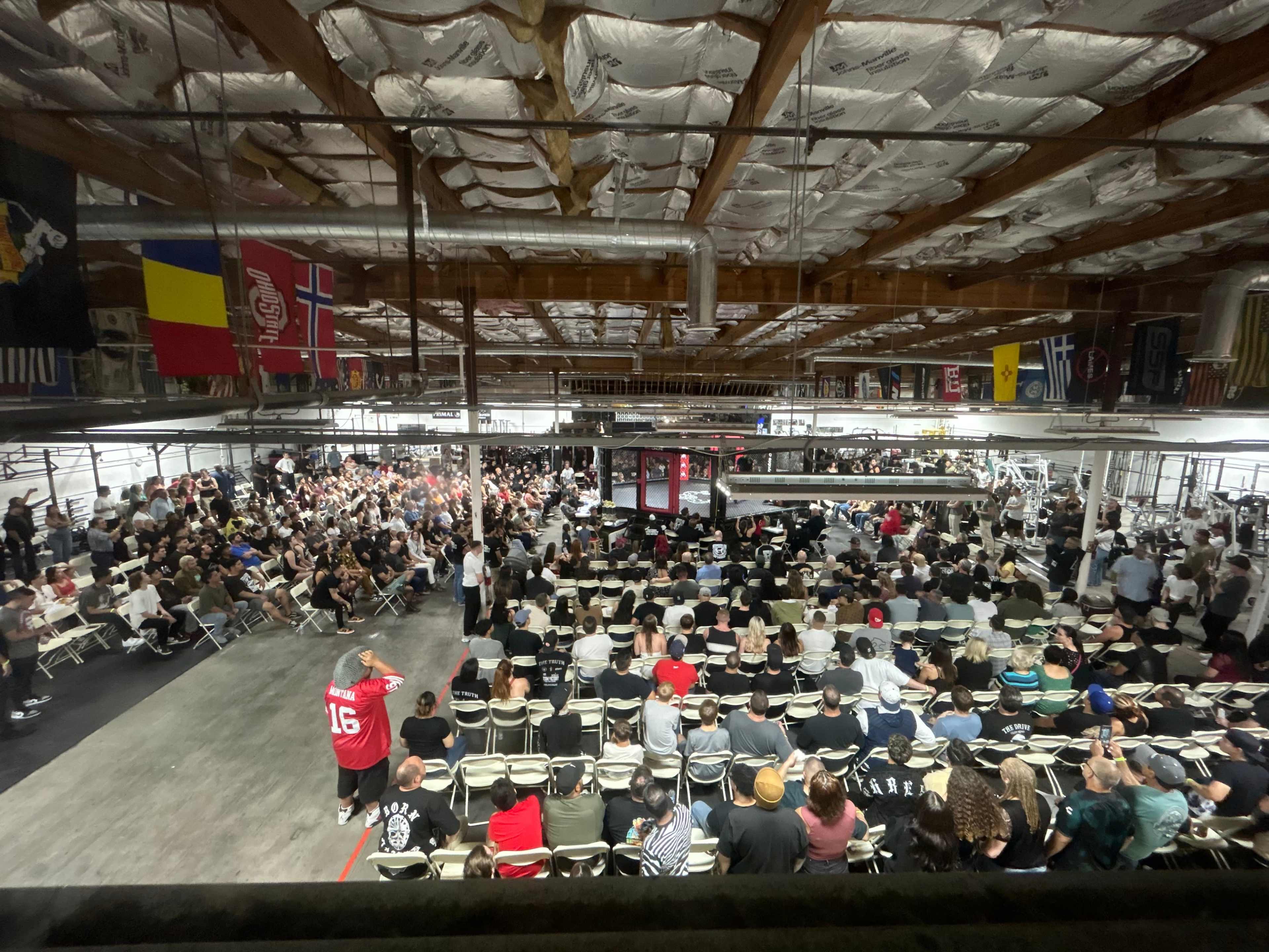 A large crowd gathers in an industrial venue filled with seating, while various flags hang from the ceiling.