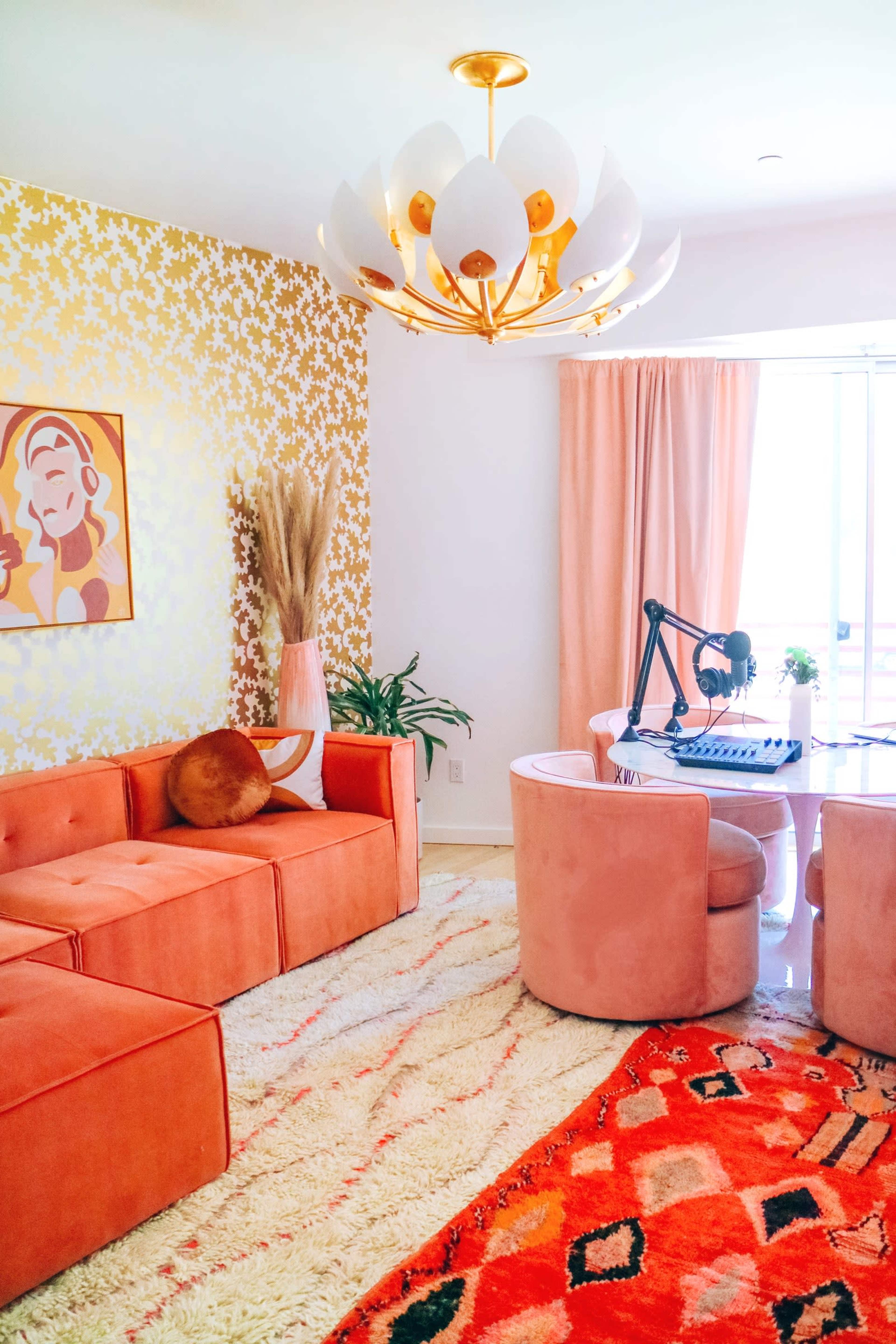 A bright, modern workspace featuring a round table with a microphone and a vibrant rug, complemented by a pink couch and gold accents.