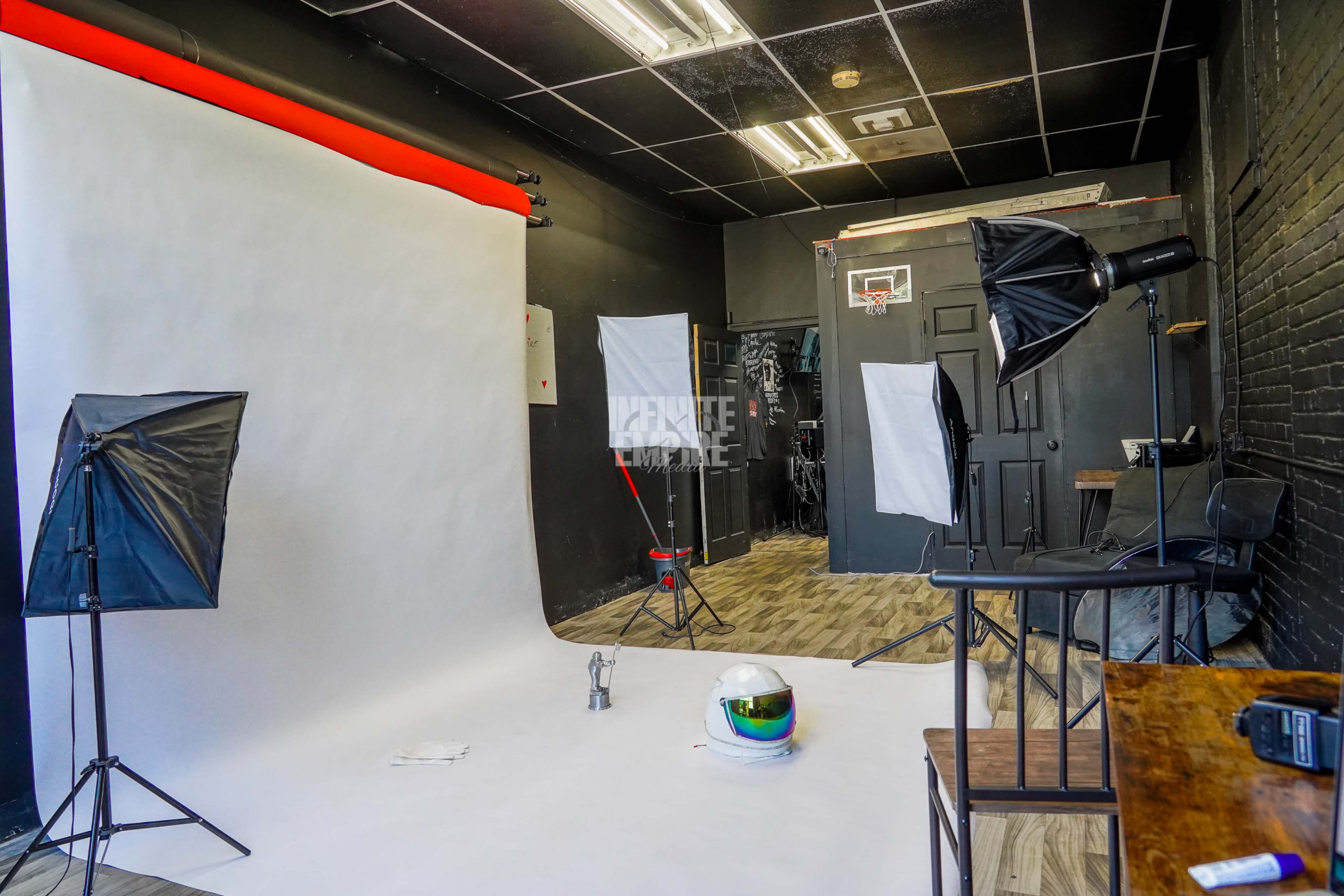 The image depicts a photography studio featuring a white backdrop, studio lights, a chair, and various equipment arranged against a black brick wall.