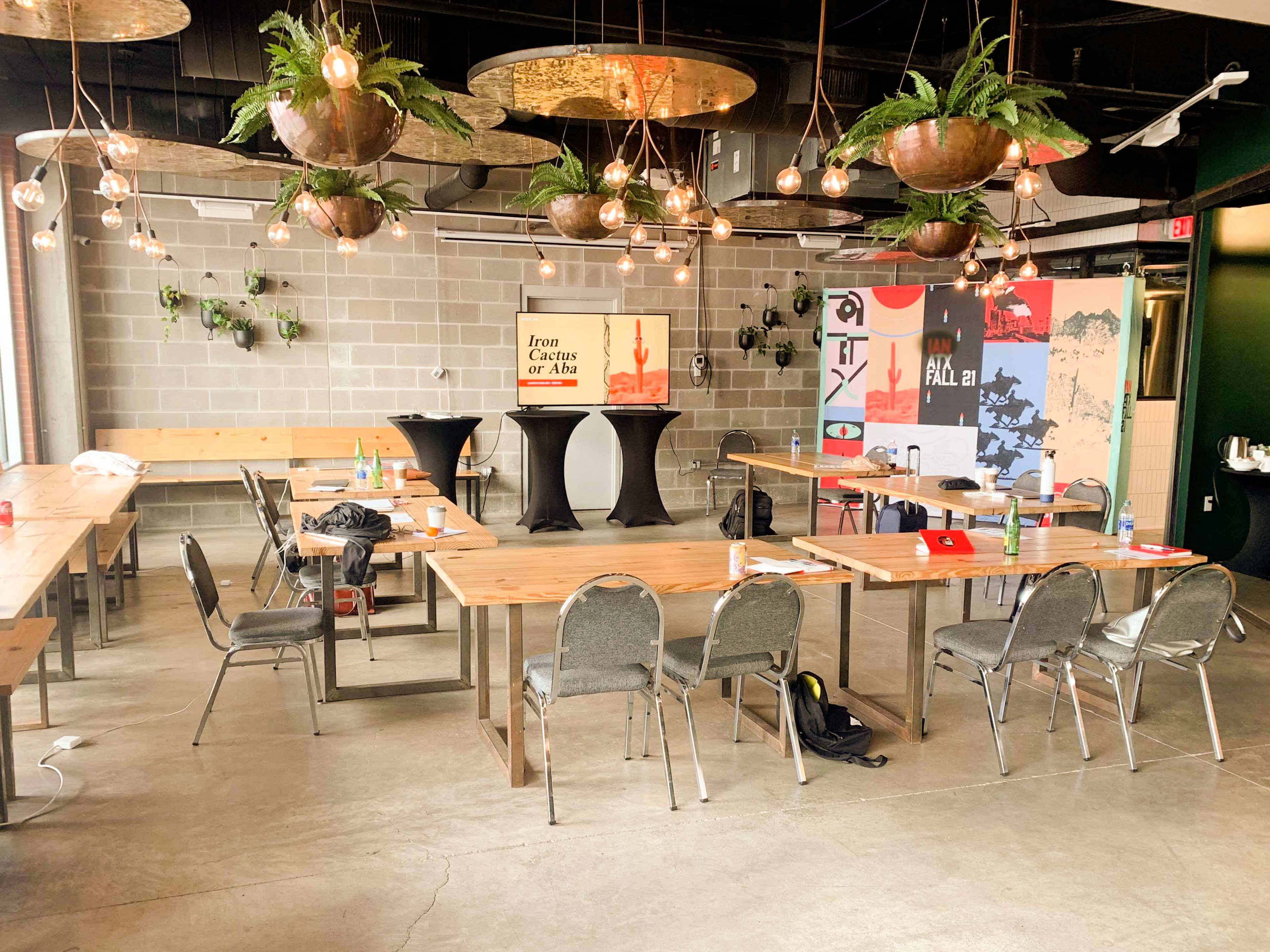 The image shows a modern event space with wooden tables, chairs arranged around the room, and decorative pendant lights hanging above, featuring a podium and a backdrop with vibrant graphics.