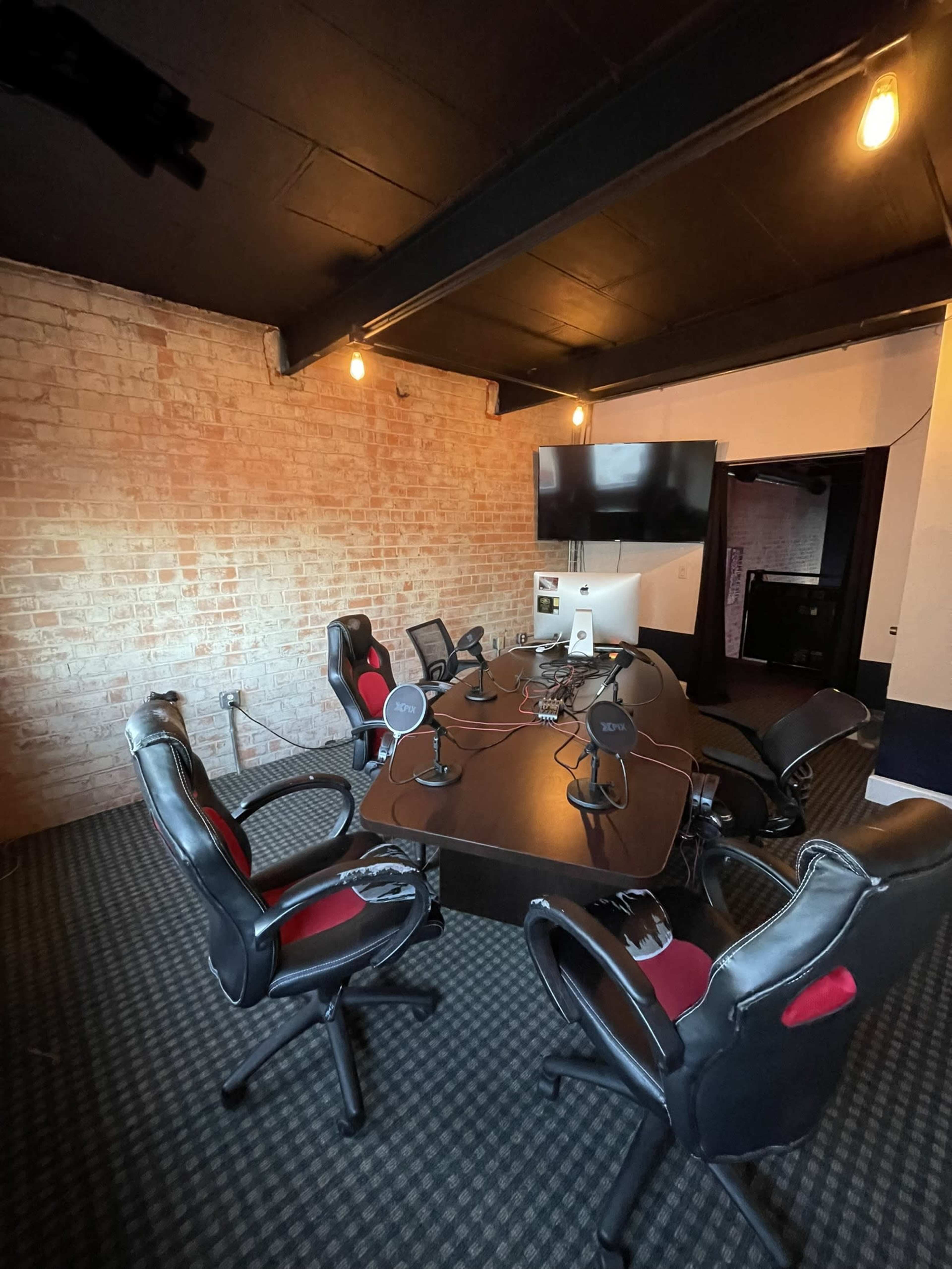 An office room with a large conference table surrounded by several black and red chairs, equipped with microphones and a computer, set against a brick wall.