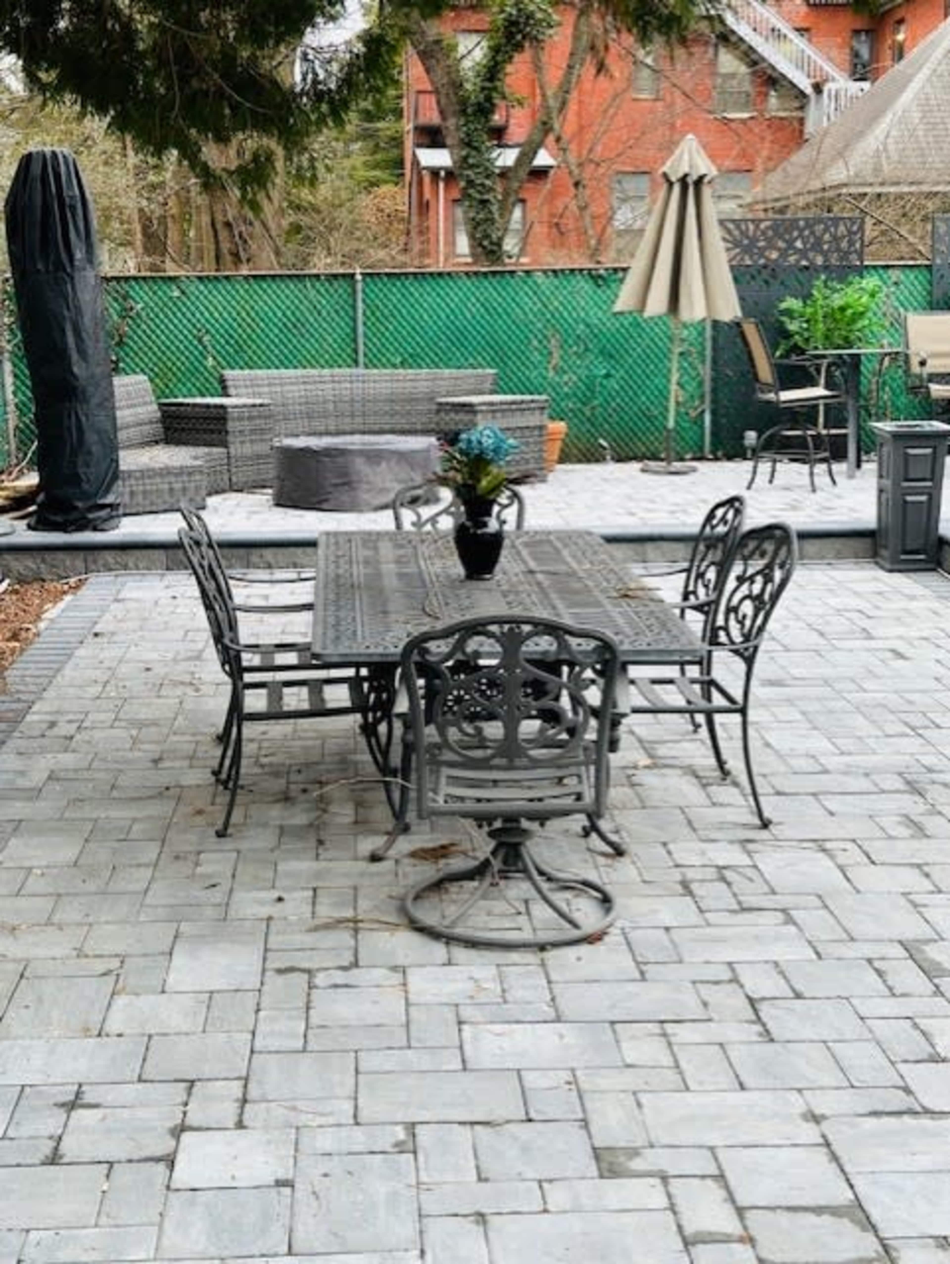 A patio area features a metal dining table surrounded by five chairs, with a large umbrella and various seating arrangements in the background.