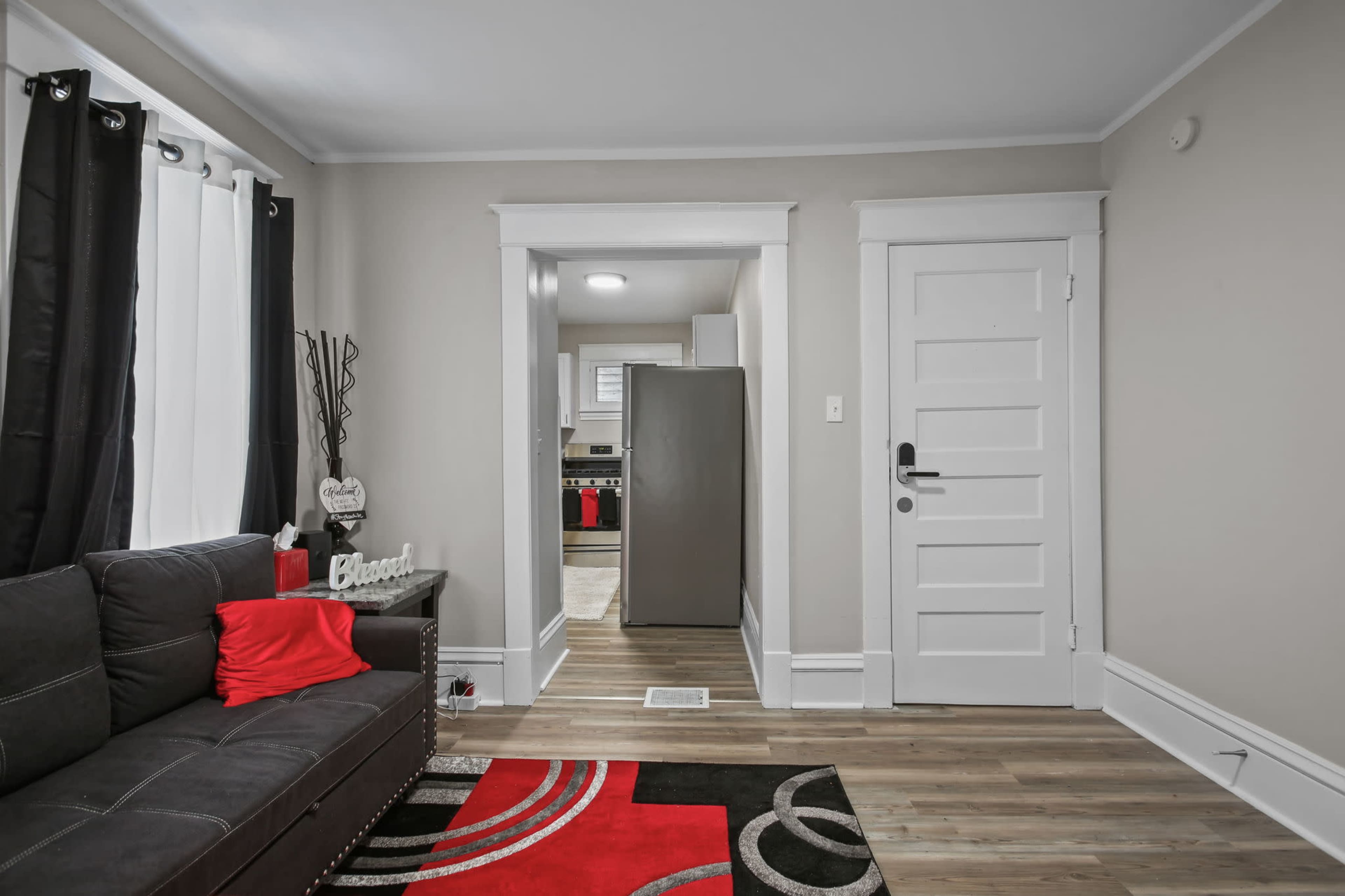 A living room with a black couch, a red accent pillow, and a view of a kitchen through an open doorway.