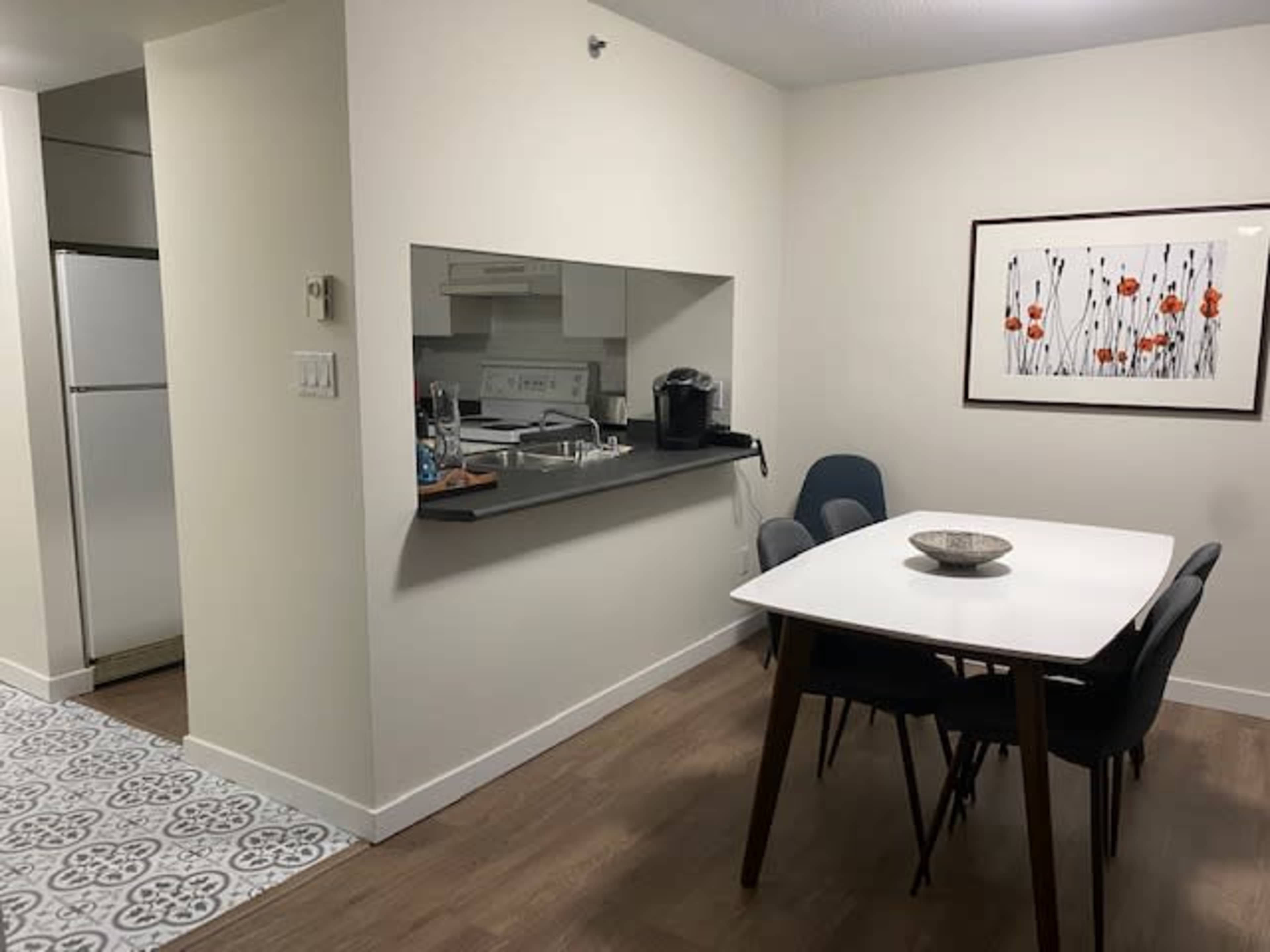 The image shows a modern dining area with a white table and dark chairs, adjacent to a kitchen area, and a decorative wall art featuring floral designs.