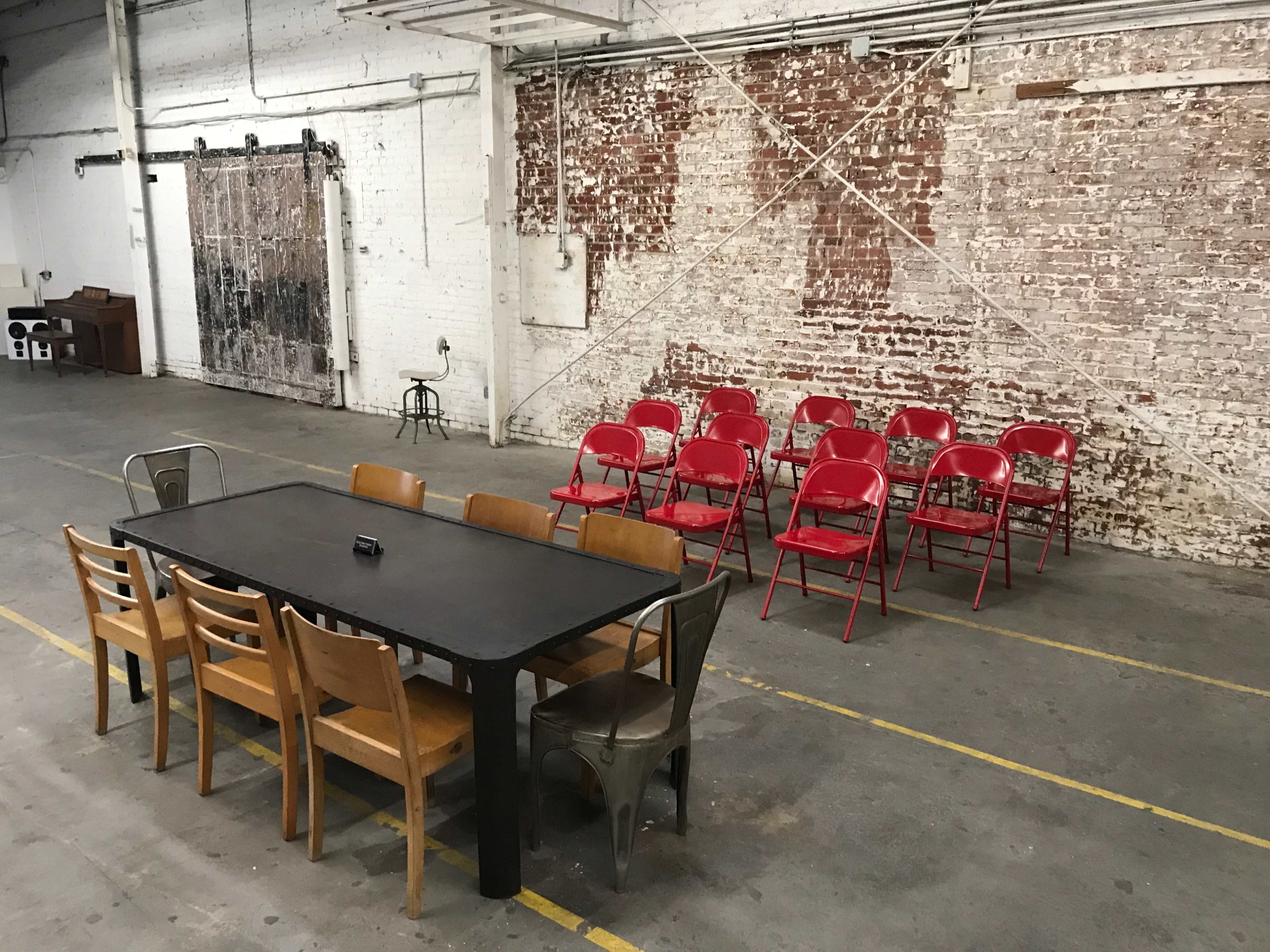 A long black table with wooden chairs is positioned in front of a row of red folding chairs against a brick wall in an industrial space.