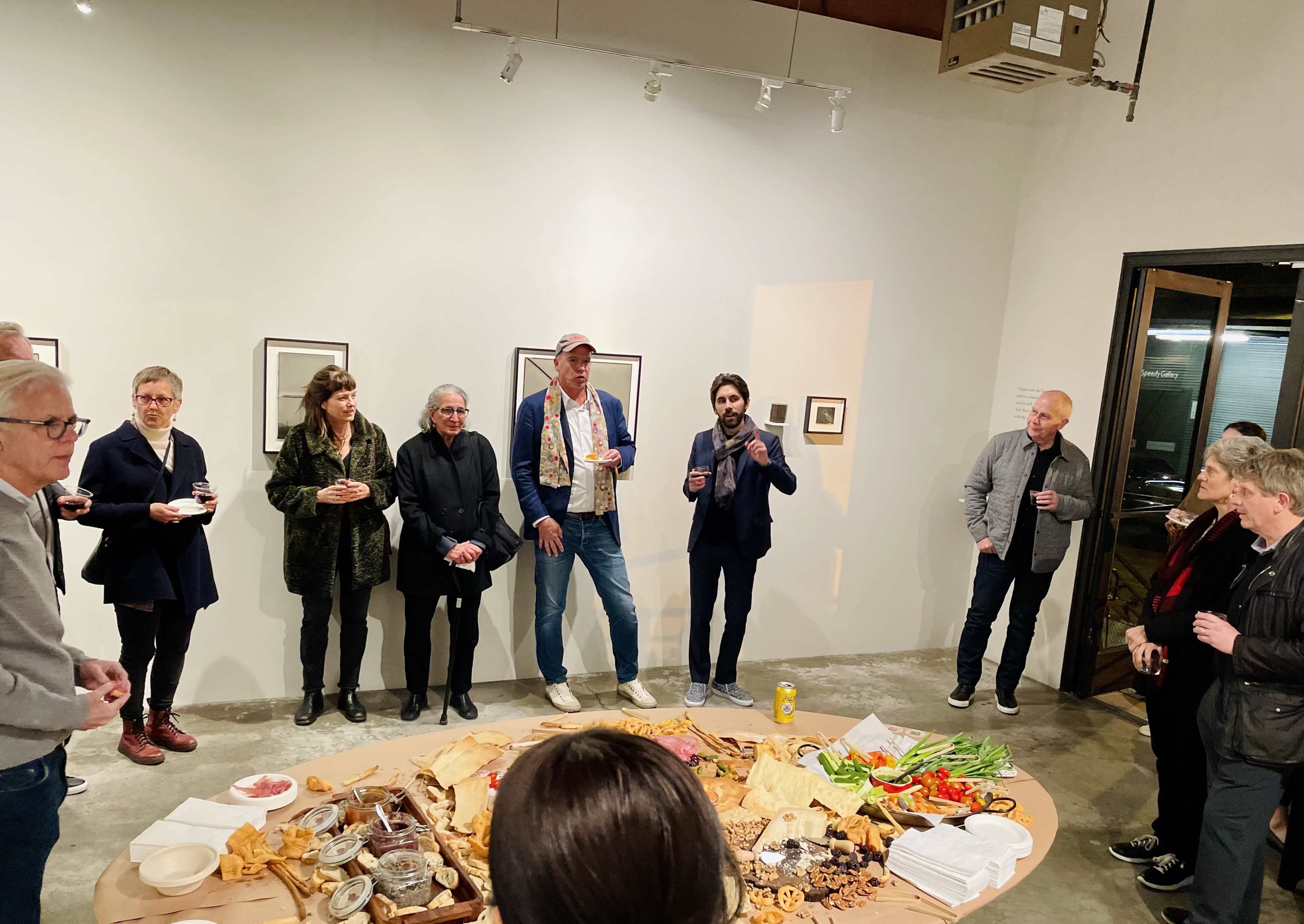 A group of people stands in an art gallery, engaged in conversation around a table filled with various food items.