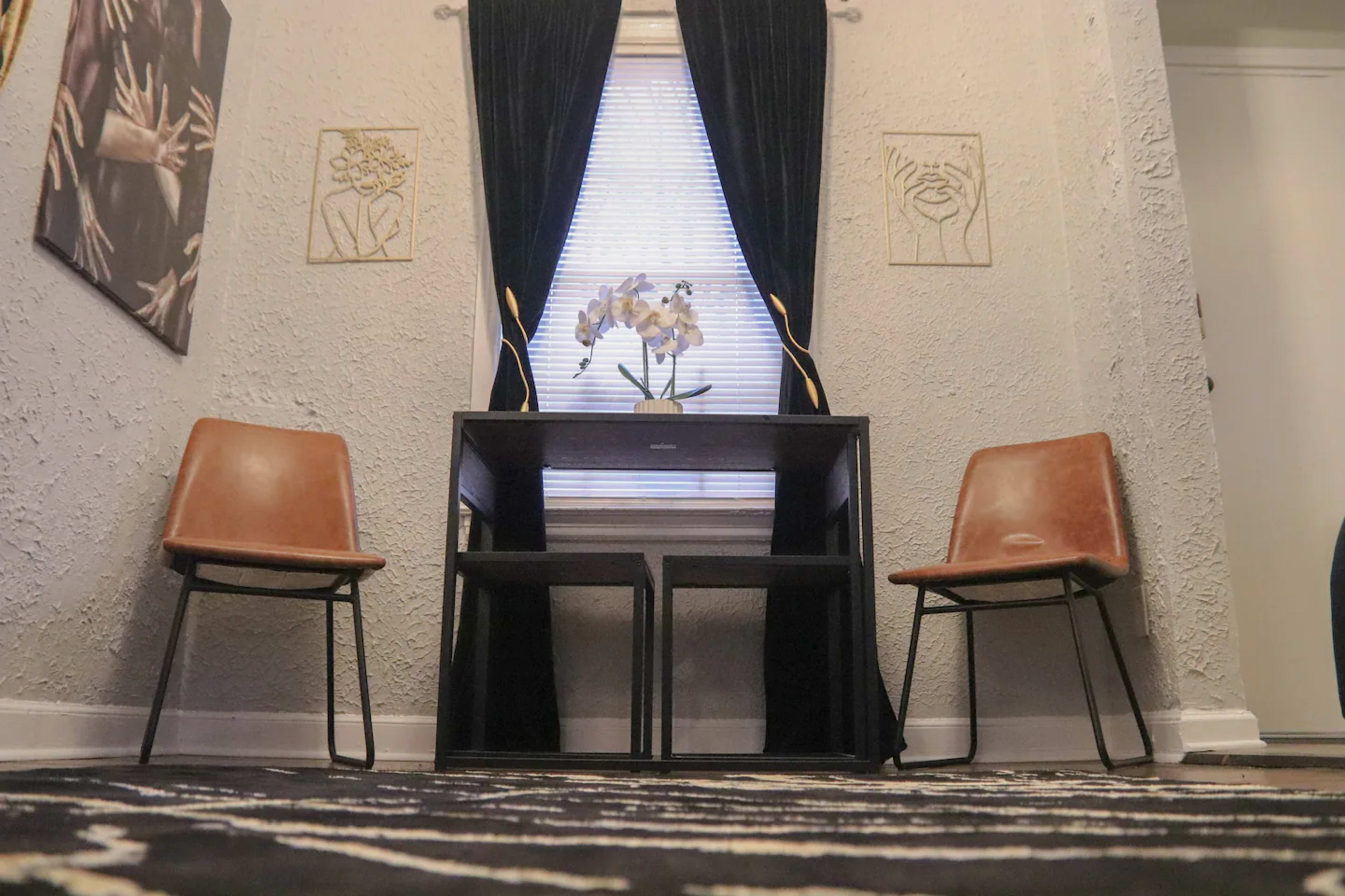 A small room with a desk and two chairs facing each other, adorned with wall art and a window draped with black curtains.