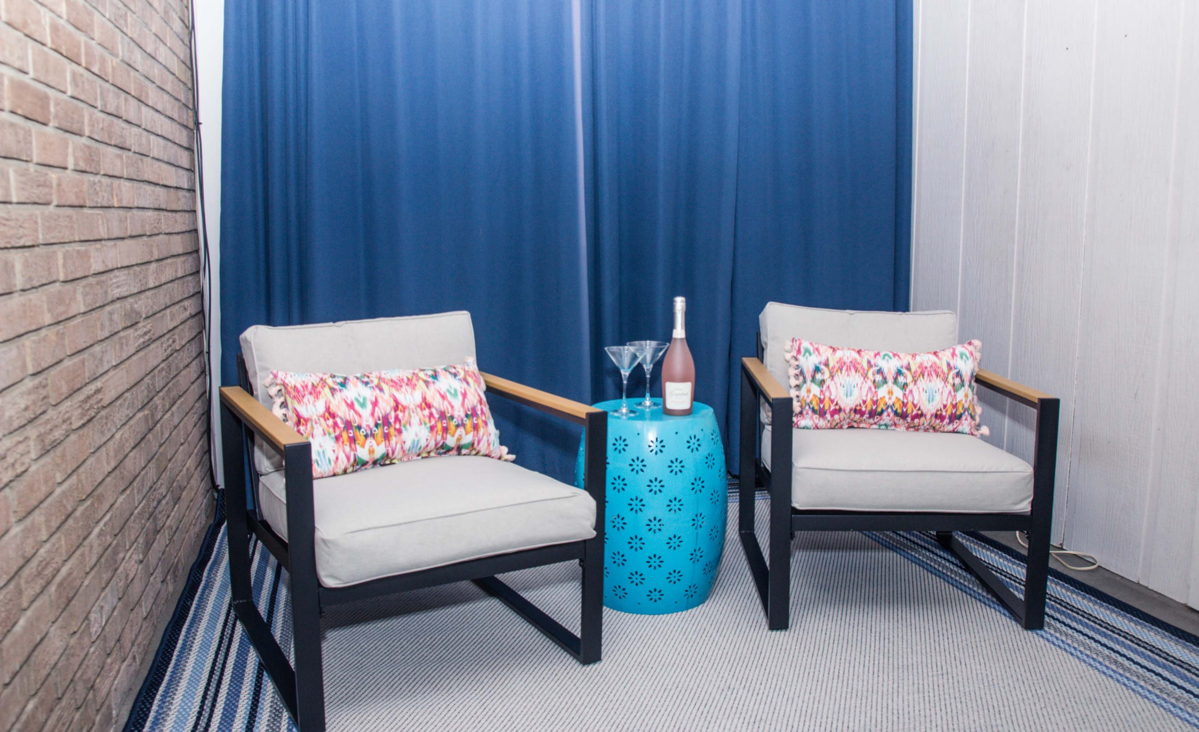 A cozy seating area features two gray armchairs with colorful cushions beside a blue decorative table, set against a backdrop of blue curtains and a brick wall.