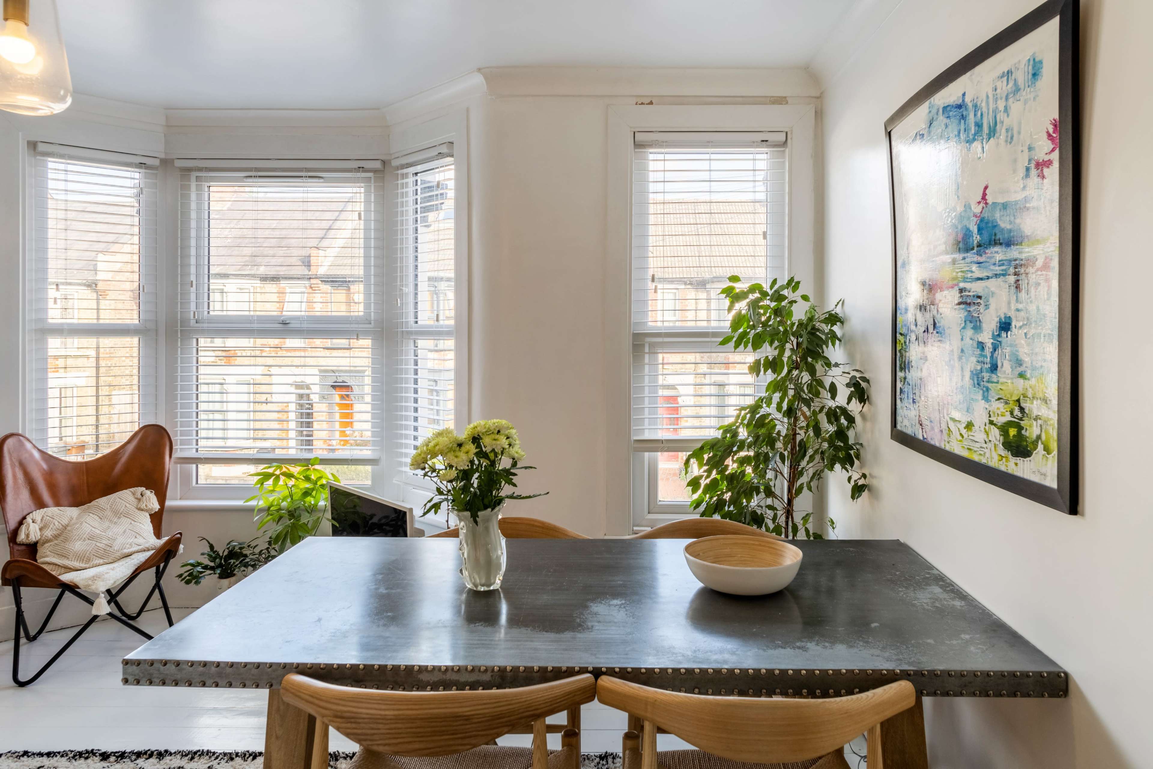A bright dining area features a metal table, a vase of flowers, a wooden bowl, and a large painting, accompanied by plants and a comfortable chair.