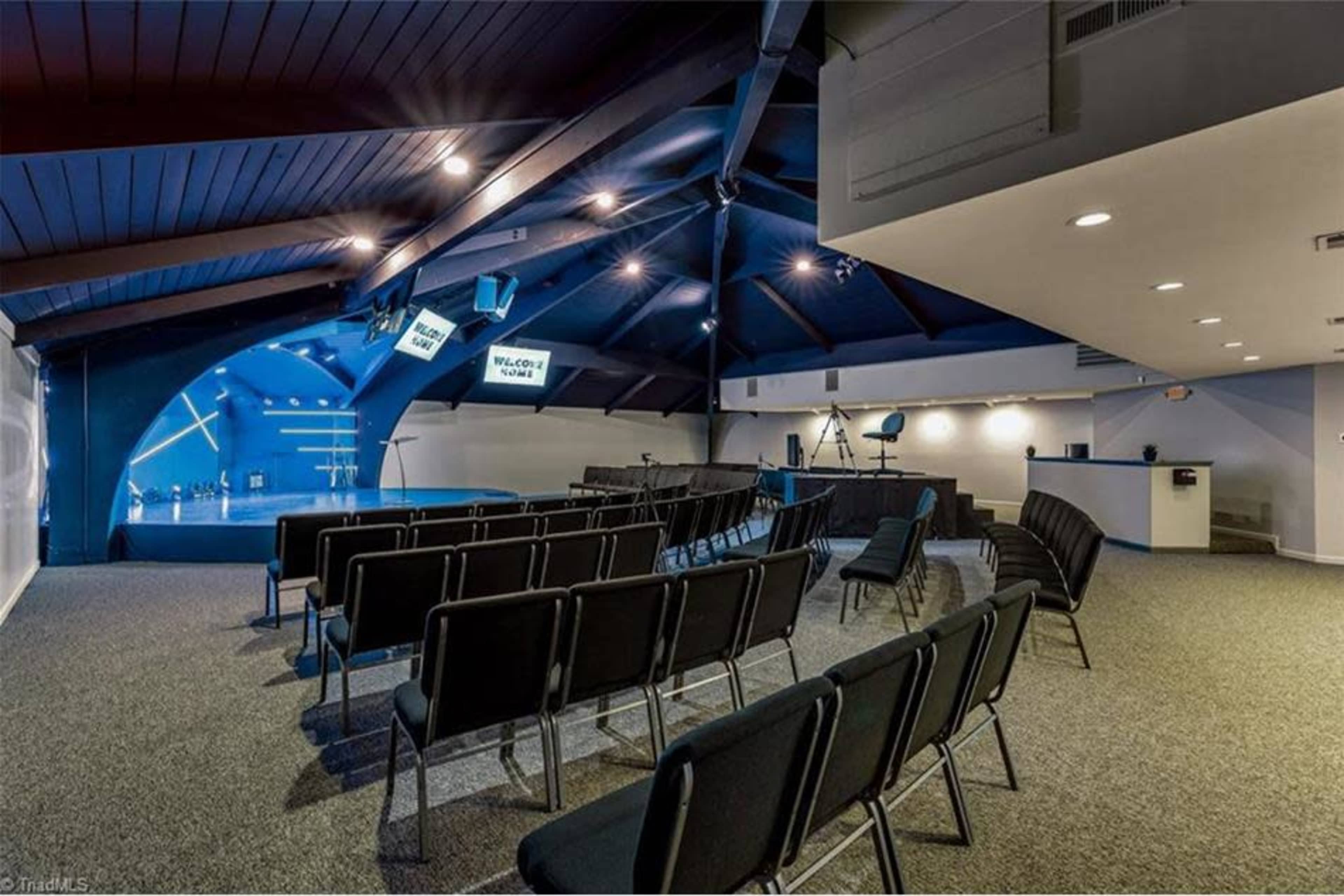 A spacious event room with black chairs arranged in rows facing a stage, illuminated by overhead lights and featuring a large blue wall.