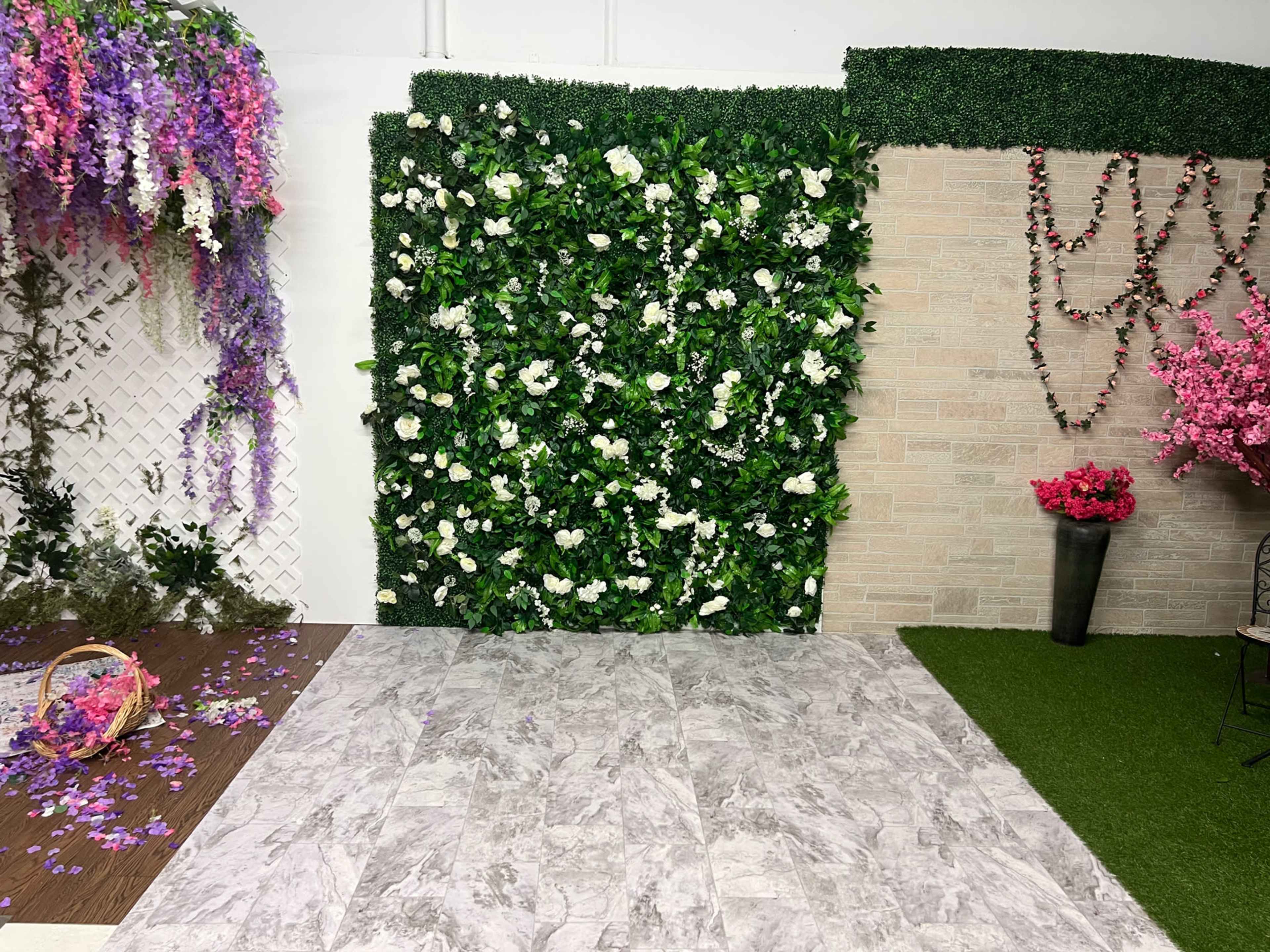 The scene features a decorated indoor space with a green plant wall, floral accents, and a mix of natural and textured flooring surfaces.