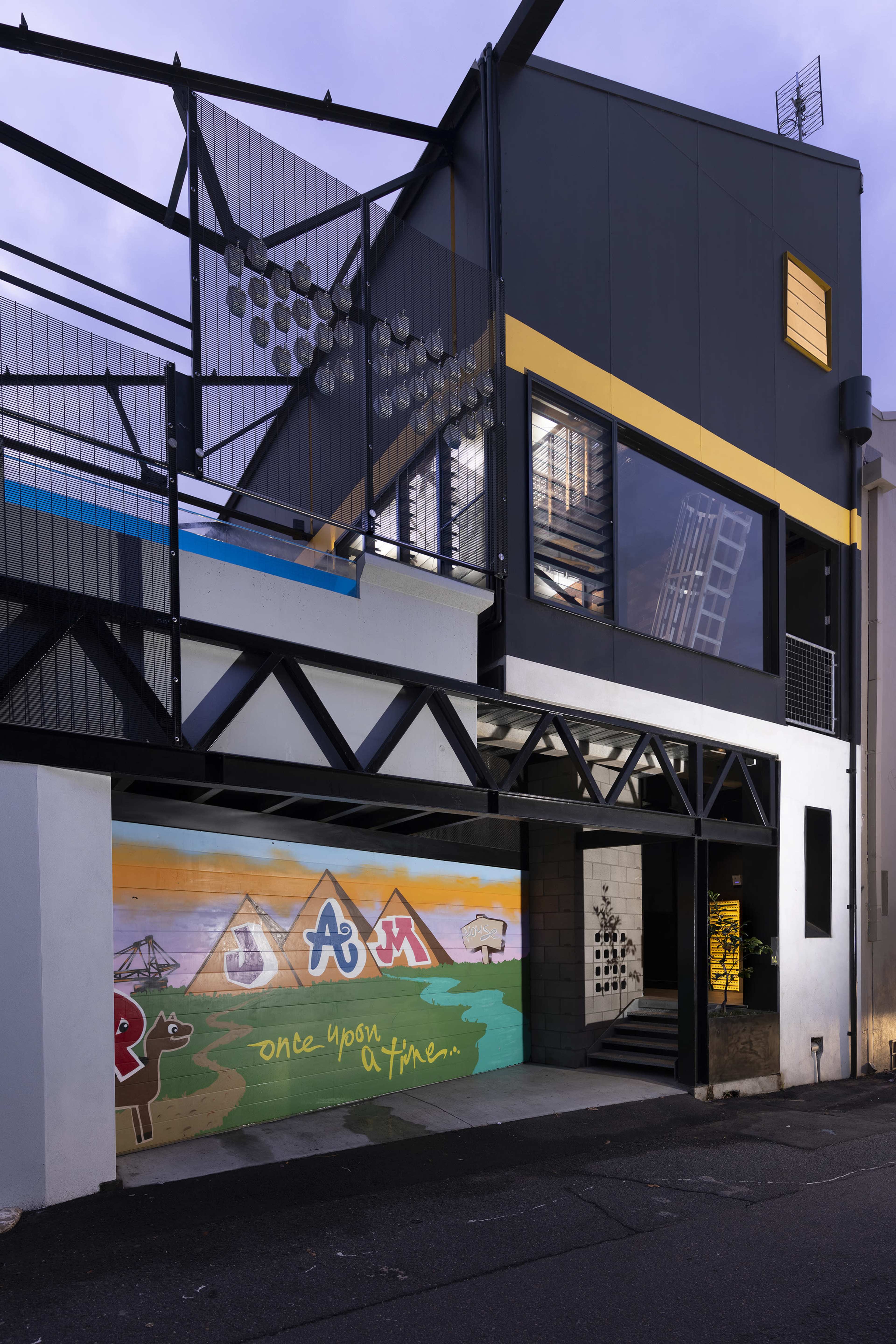 A modern building with a vibrant mural featuring mountains and the word "JAM" on one wall, alongside an industrial-style structure with large windows and metal balconies.