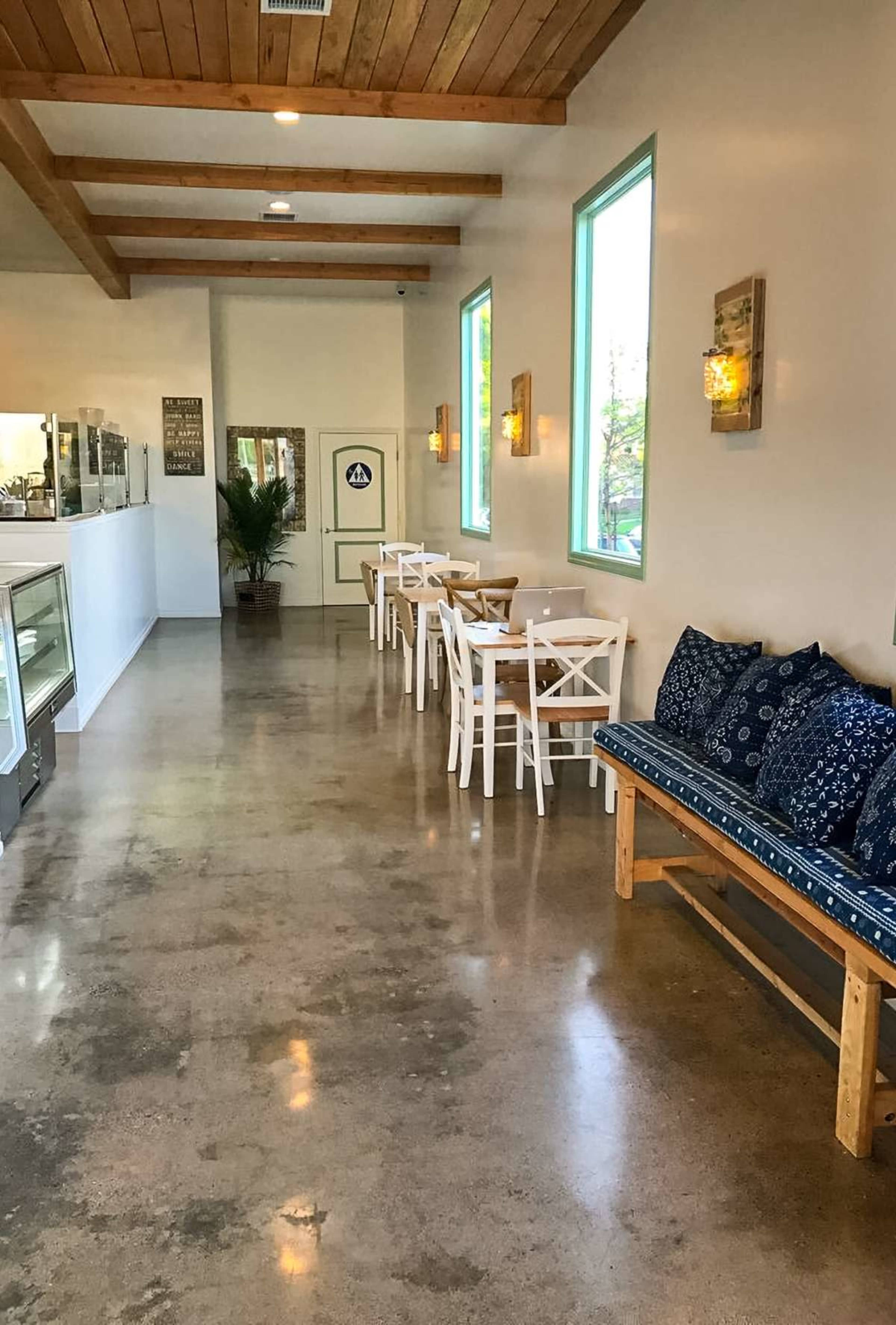 The image shows a modern café interior with concrete flooring, wooden beams, and a row of white tables and chairs along one side, complemented by a blue cushioned bench against the wall.