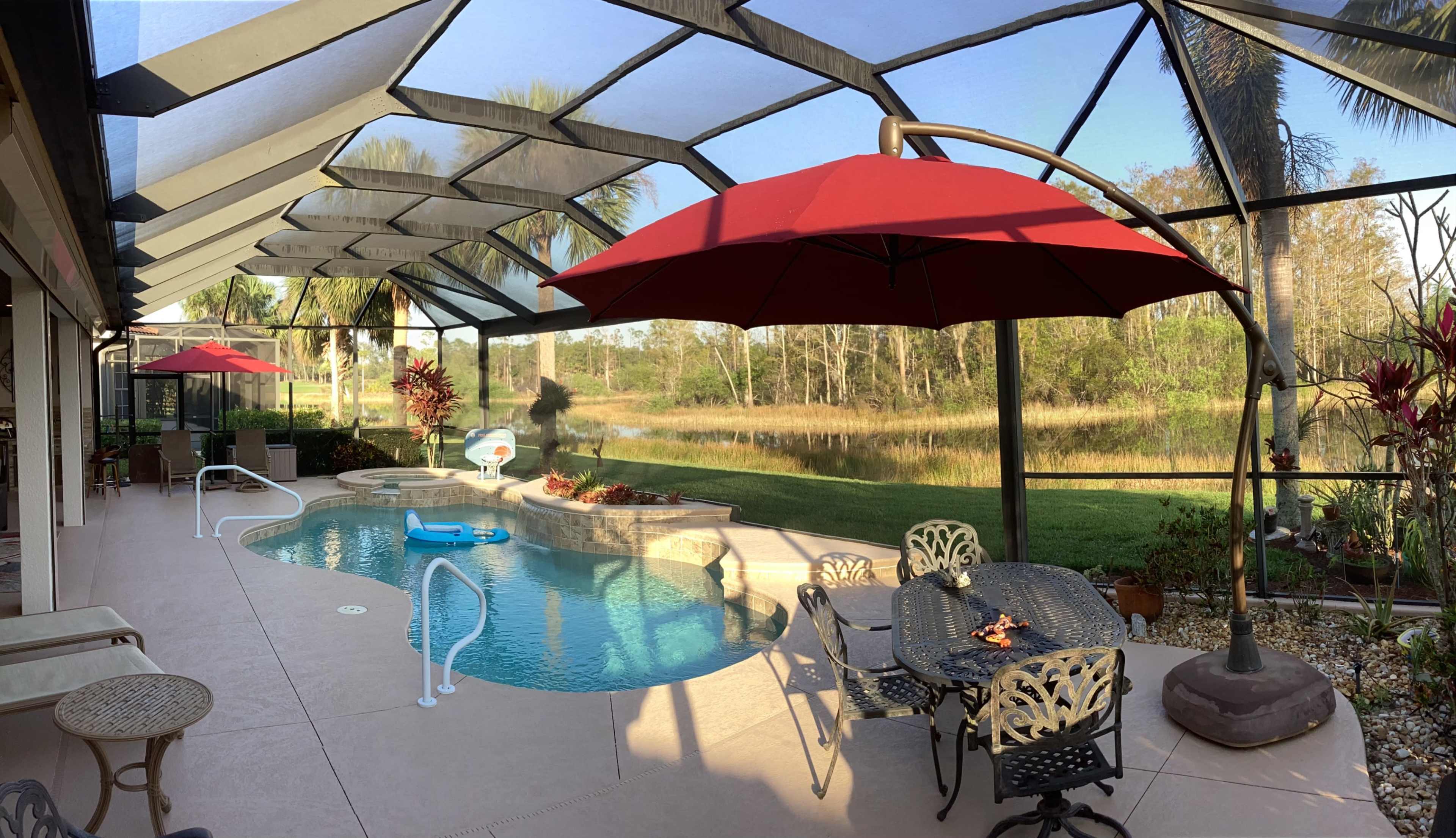A screened pool area features a swimming pool, a table with chairs, and red umbrellas overlooking a grassy area and water in the background.