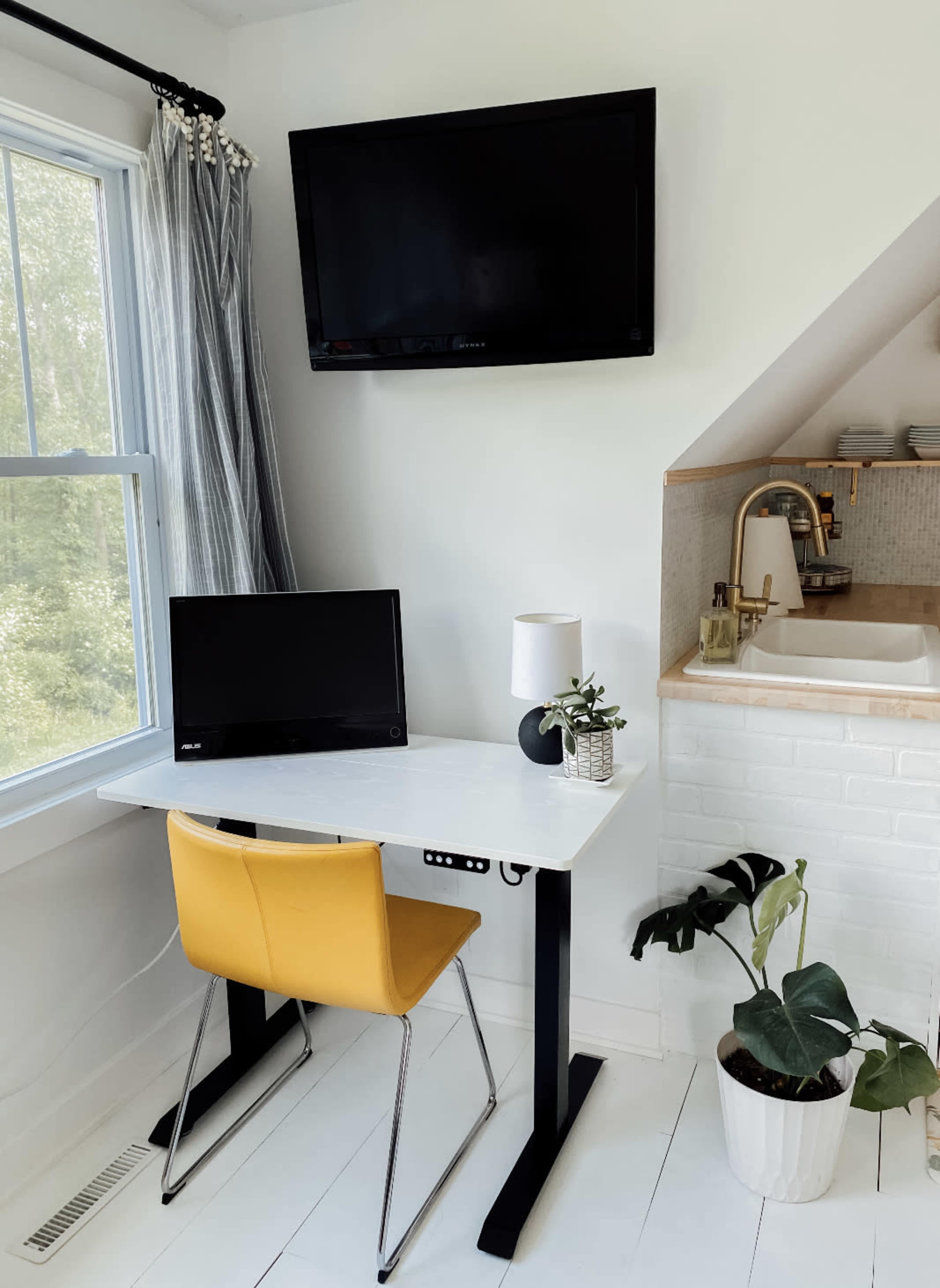 A modern workspace features a white desk with a monitor, a yellow chair, and a potted plant, set against a backdrop of a television and a kitchen sink in a bright, minimalist room.