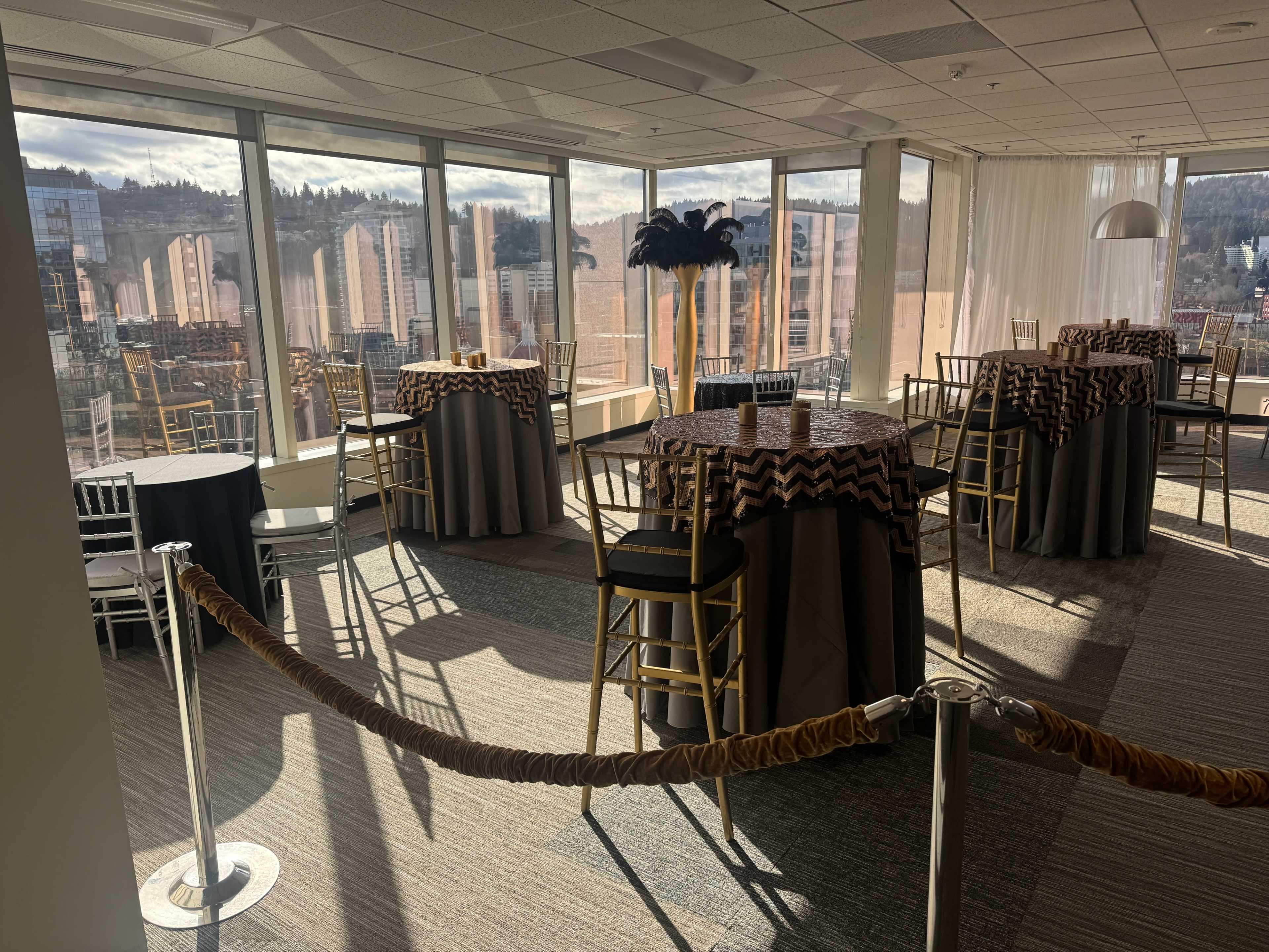 A spacious event room features several tall tables with chevron-patterned tablecloths, surrounded by large windows that offer a view of the city.