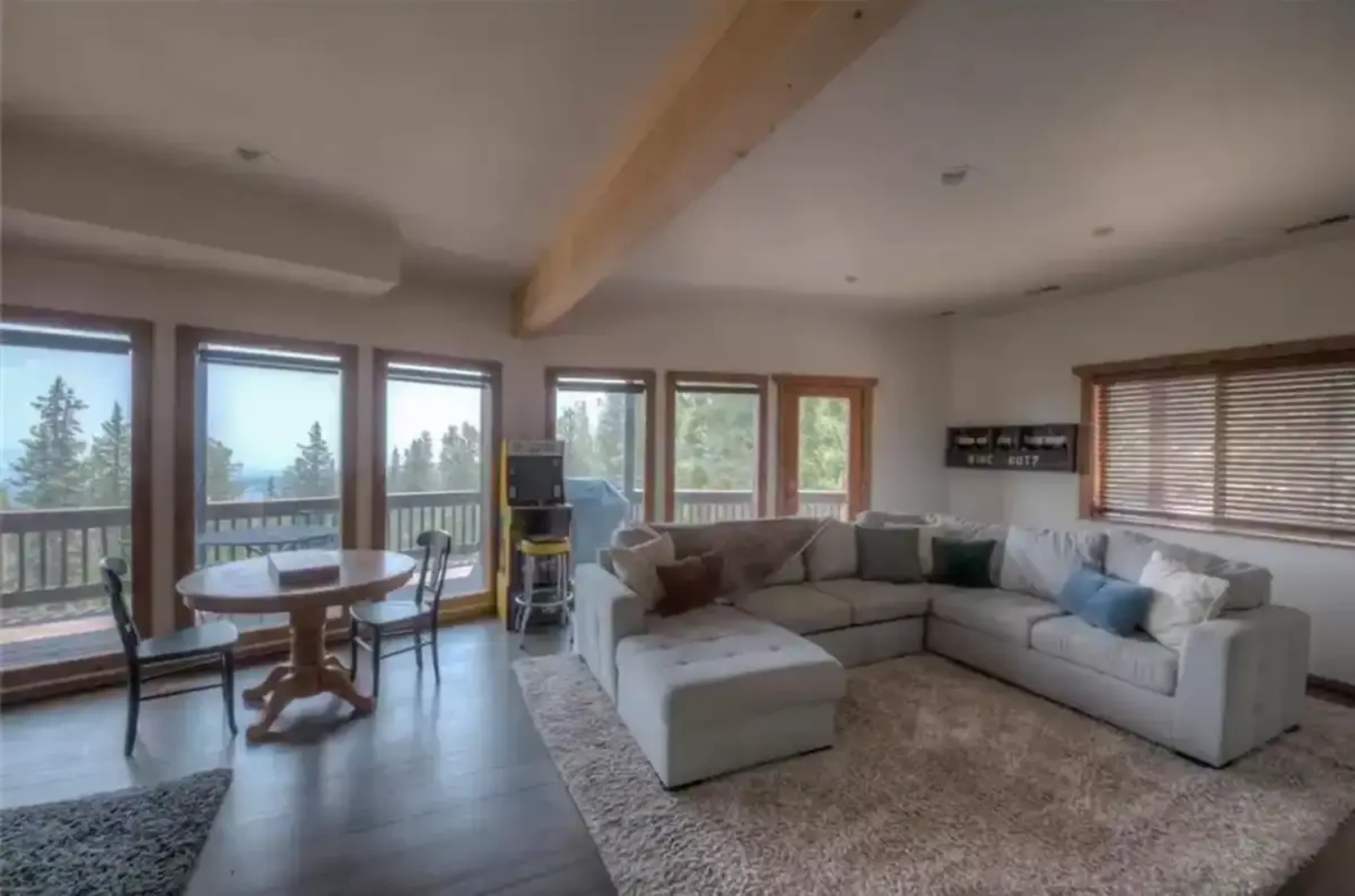 A spacious living room features a sectional sofa, a round dining table, and large windows overlooking a forested landscape.