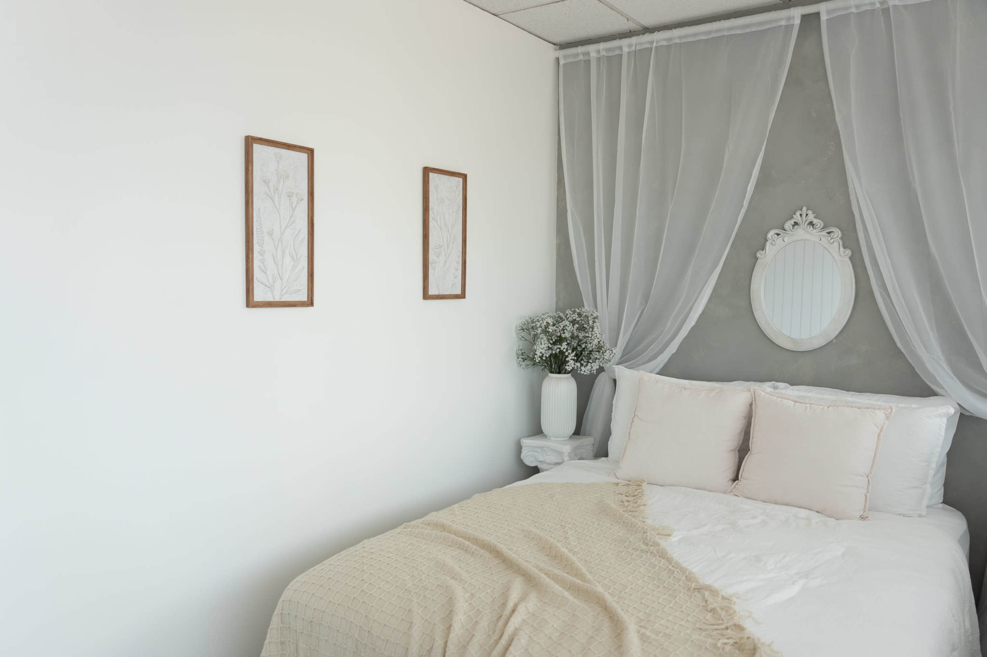 The image shows a minimalist bedroom featuring a bed with light-colored linens, two framed botanical prints on the wall, and sheer curtains draping over the bed.
