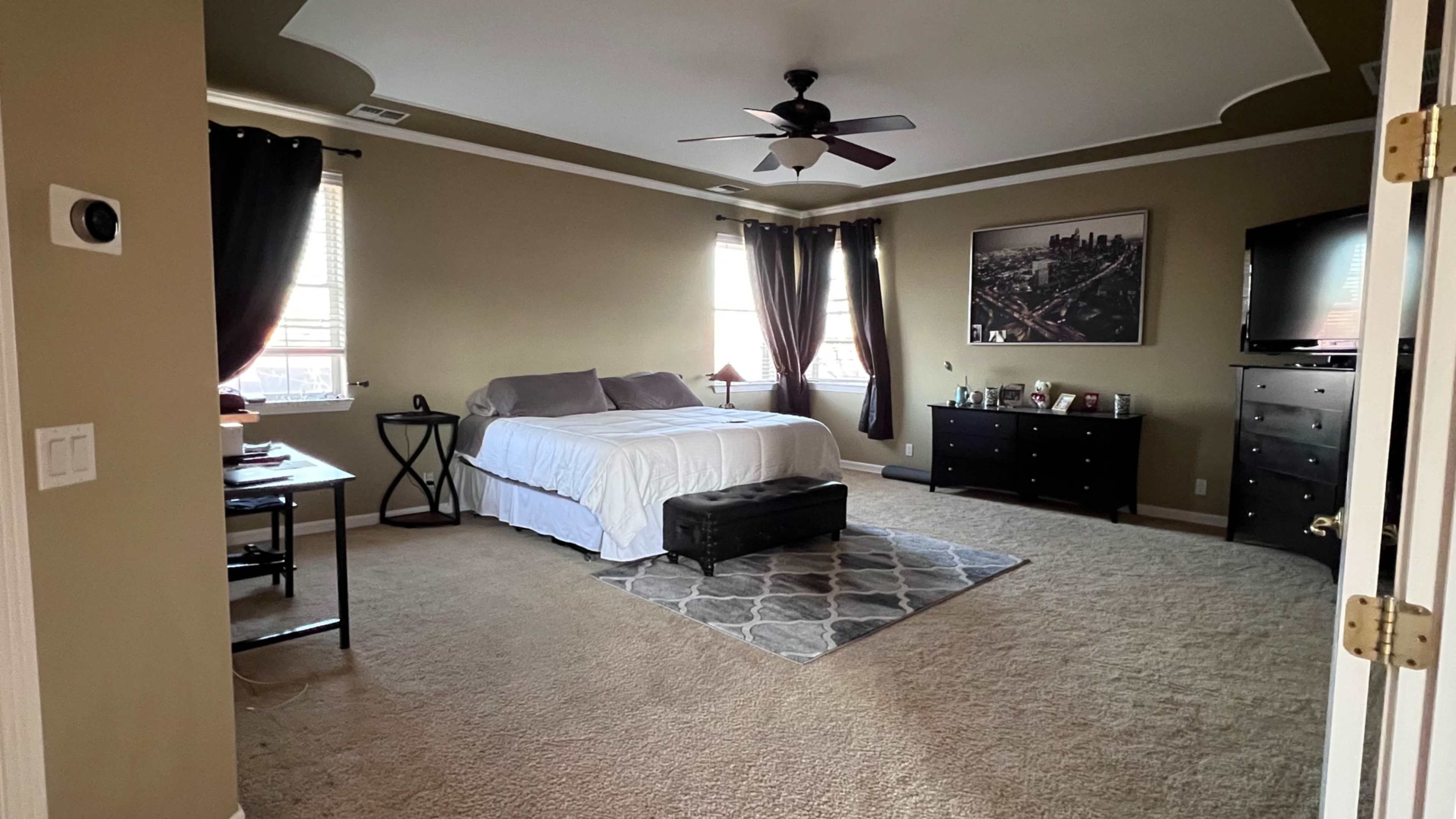 A spacious bedroom features a neatly made bed, a dark dresser, a television, and natural light coming through the windows.