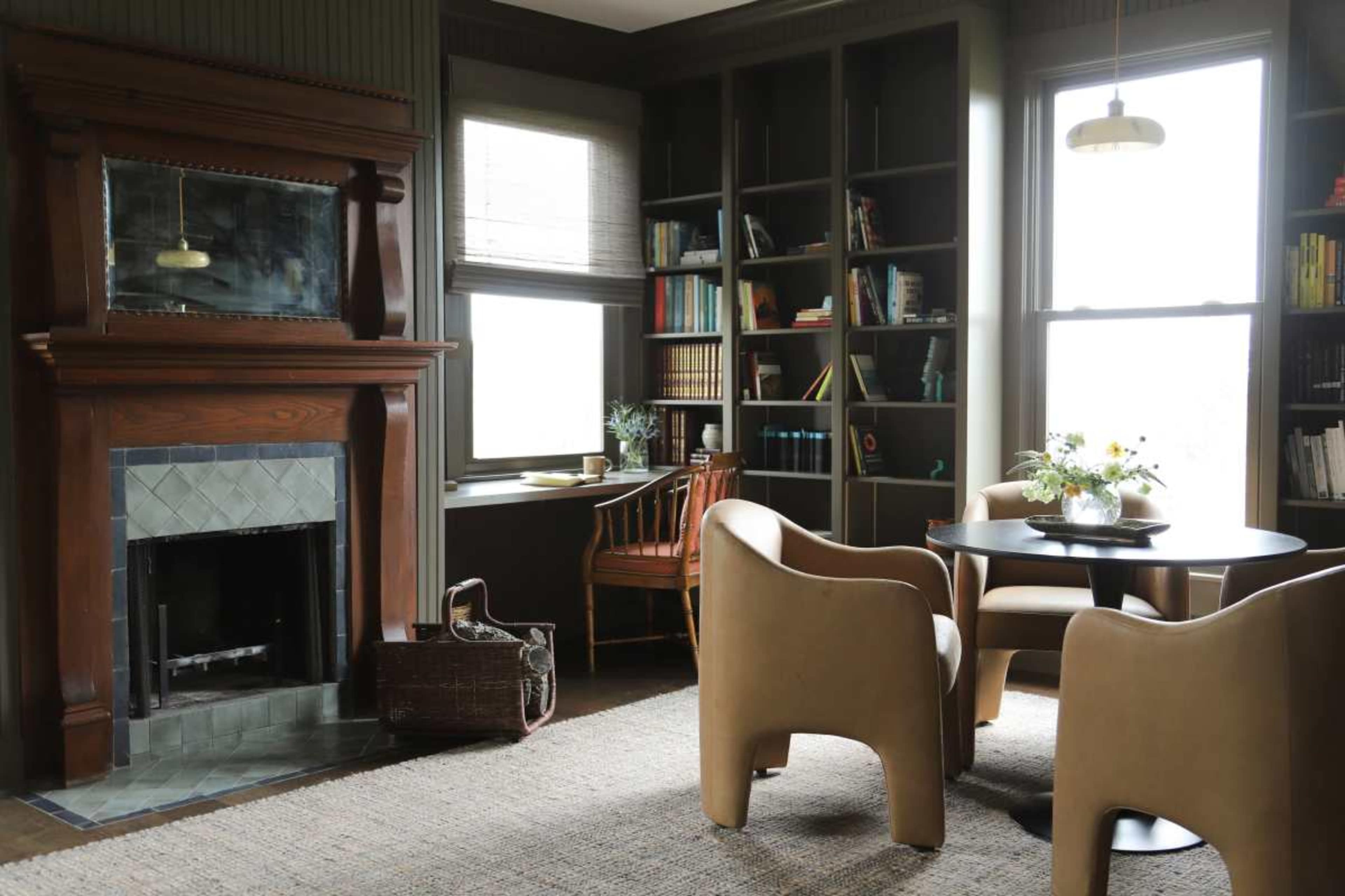 The image shows a cozy living room featuring a fireplace with an ornate mantel, a round table with two chairs, a window with bookshelves, and a basket on the floor.