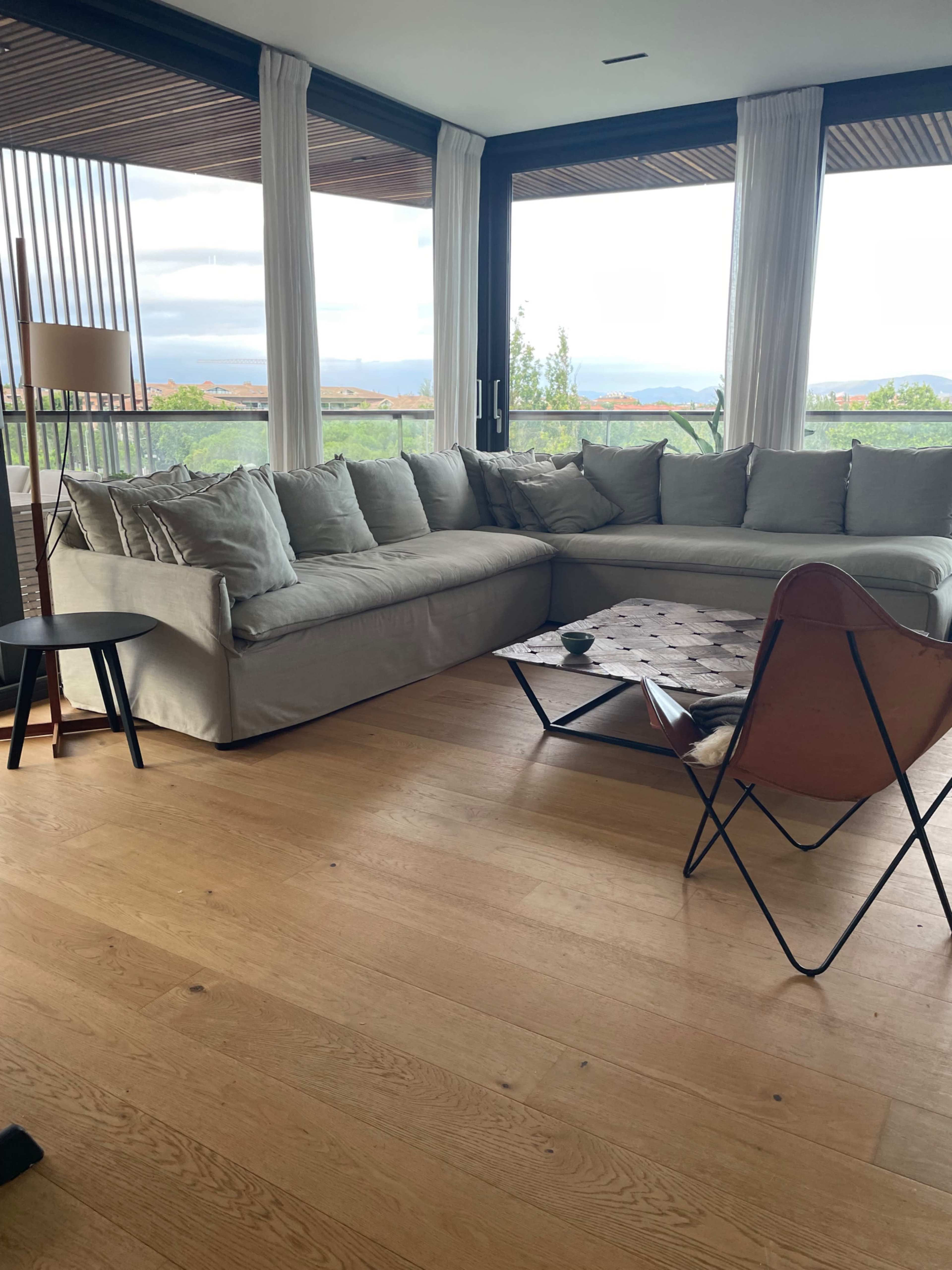 The image shows a modern living room with a large sectional couch, a wooden coffee table, a floor lamp, and large windows offering a view of the outdoors.