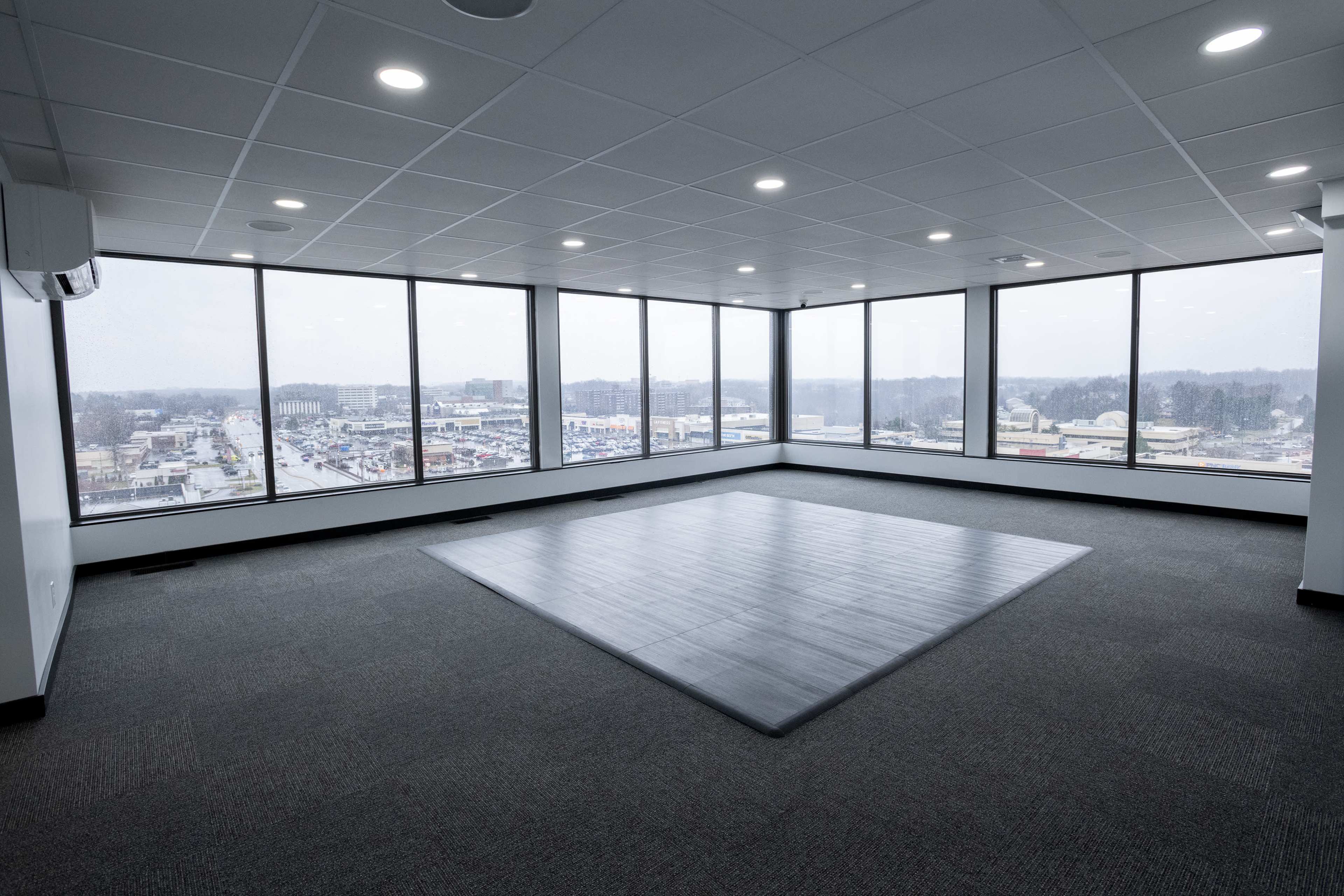 An empty office space features large windows with a view of the city and a central dark wooden platform on the carpeted floor.