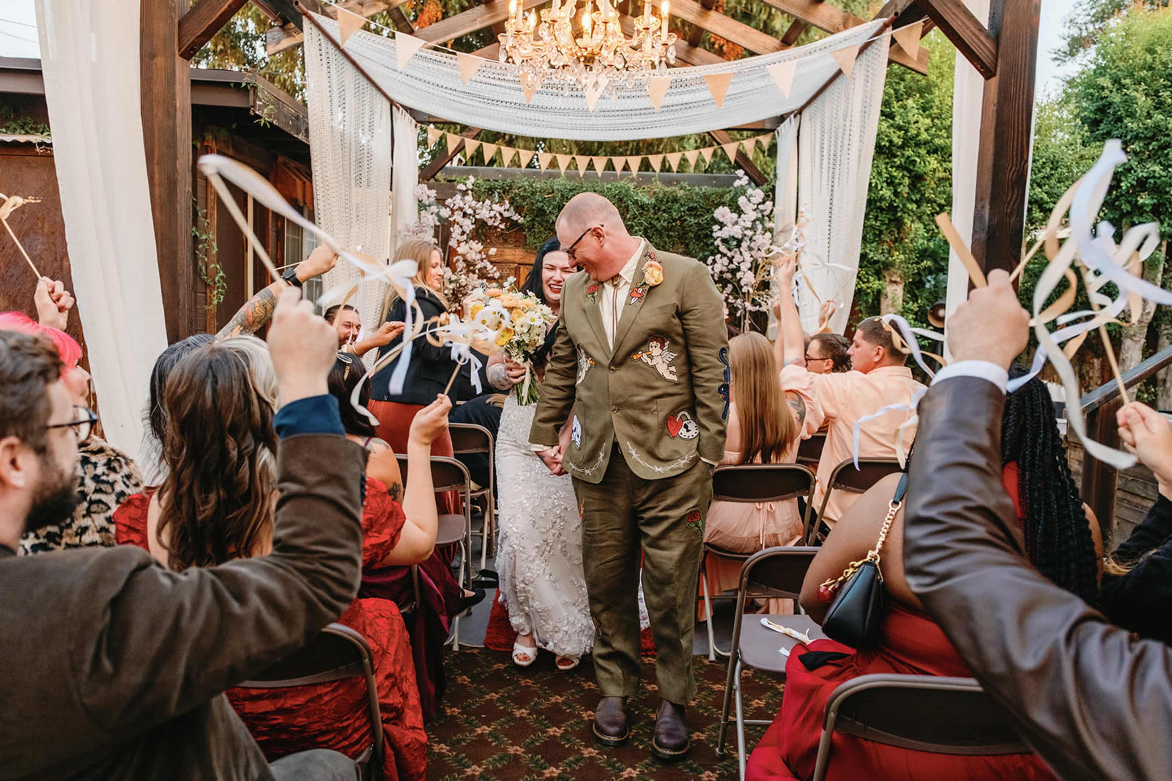 A couple walks down the aisle at a wedding ceremony, surrounded by guests holding streamers and decorated with floral arrangements.