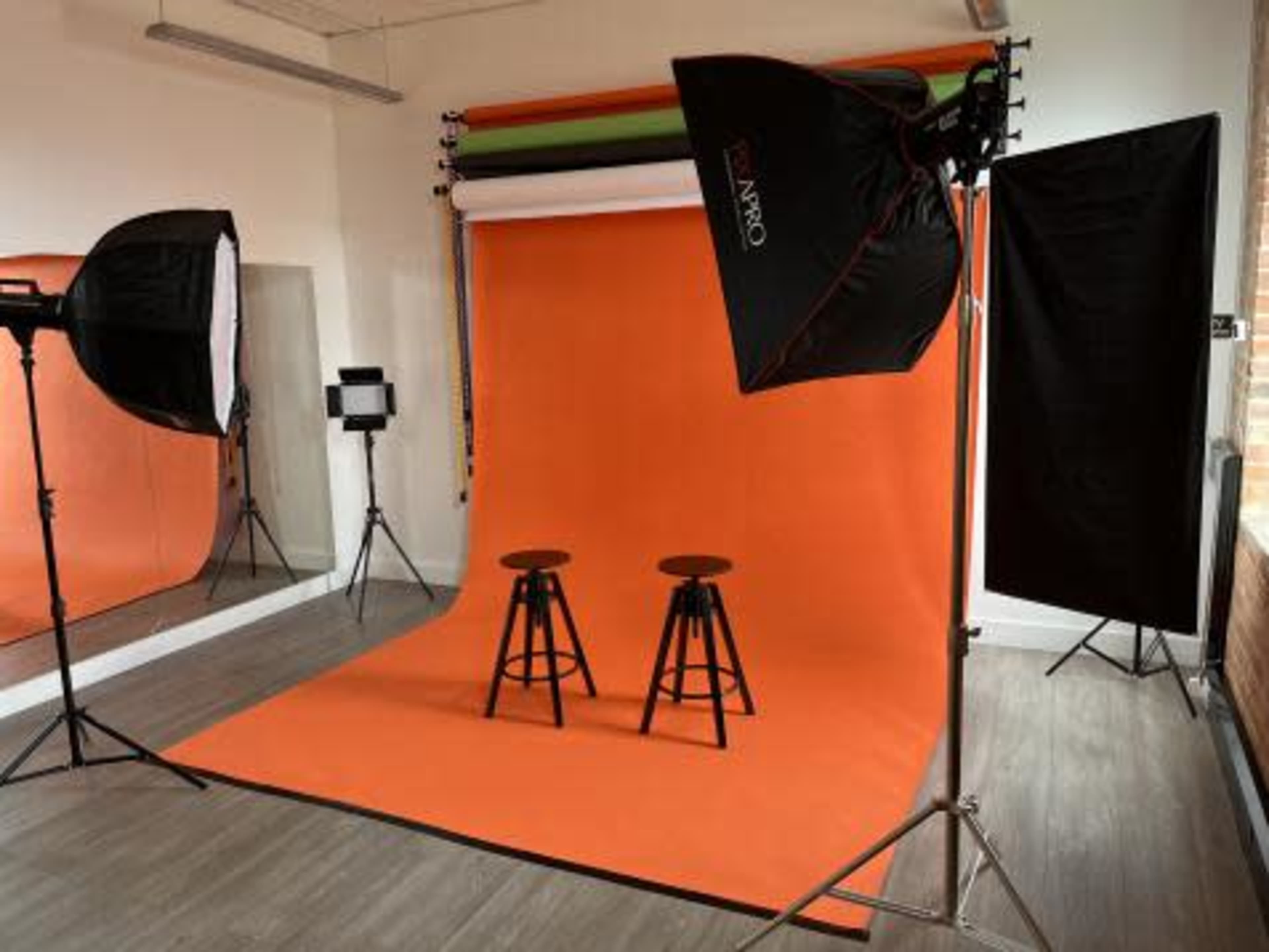 A photography studio featuring two wooden stools in front of an orange backdrop, with softbox lights positioned around the area.