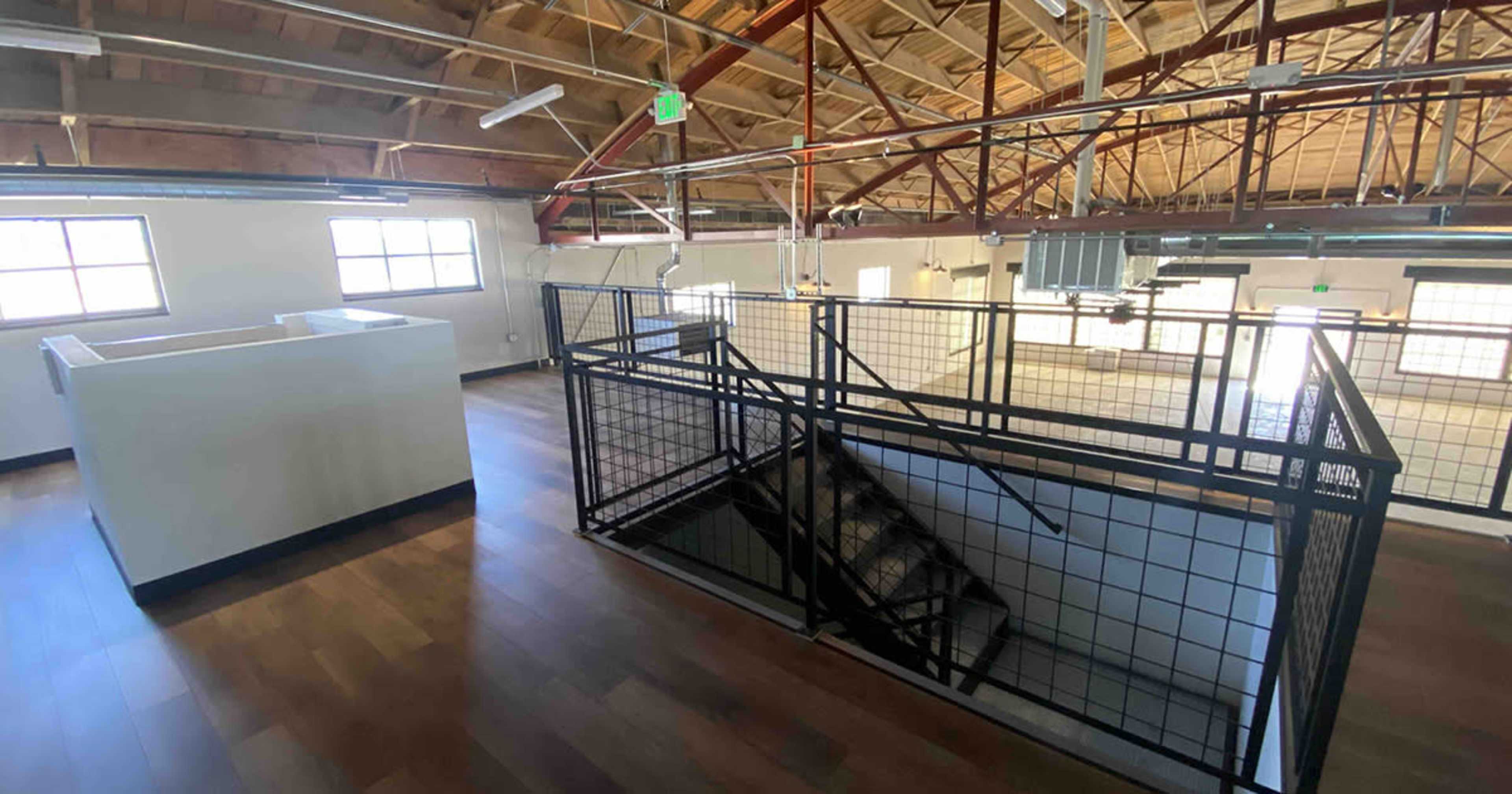 The image shows an open interior space with a high ceiling, wooden beams, and a staircase surrounded by a black metal railing.