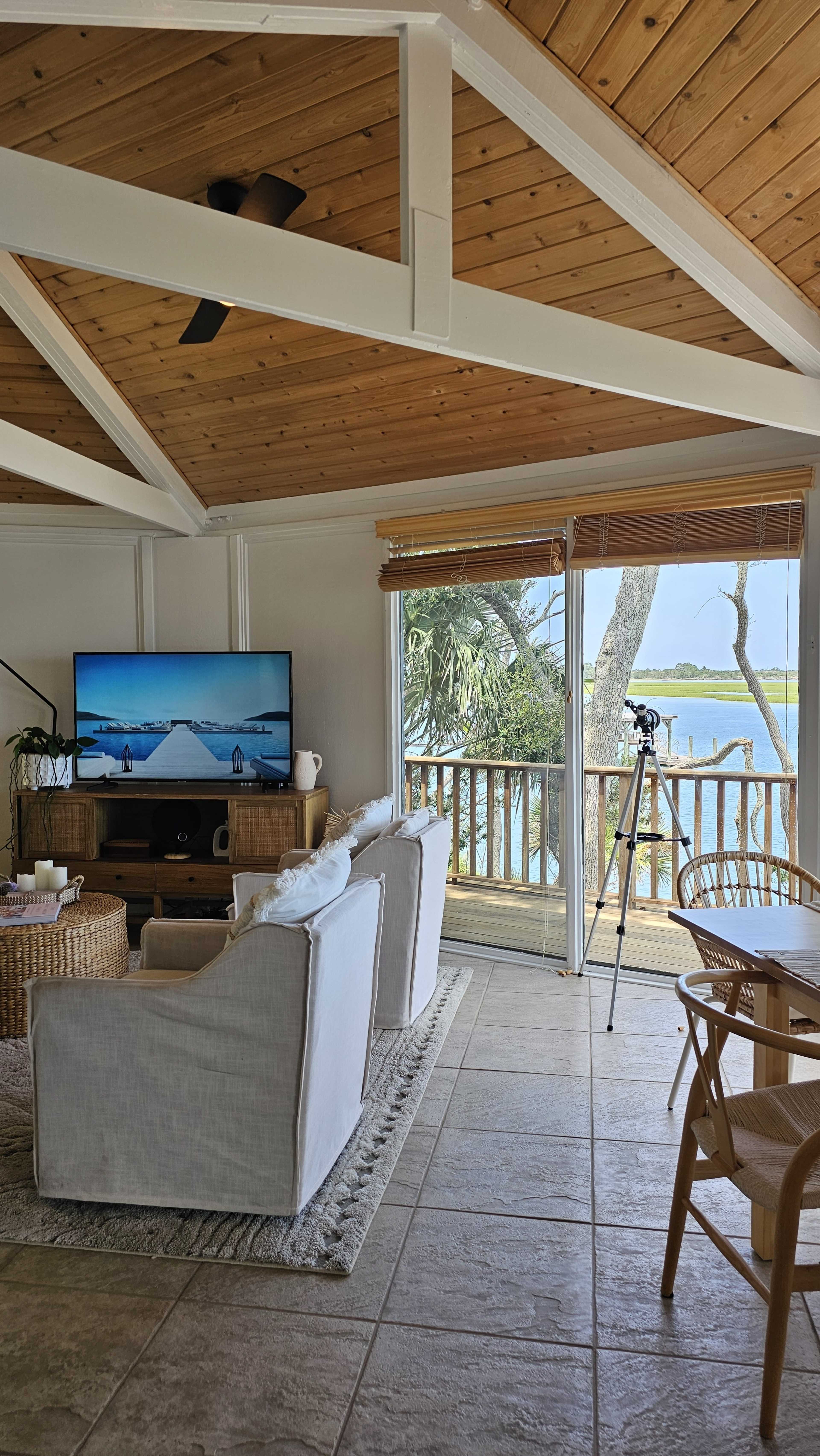A cozy living room features two white chairs facing a large television, with sliding glass doors leading to a view of a body of water and a tripod set up nearby.
