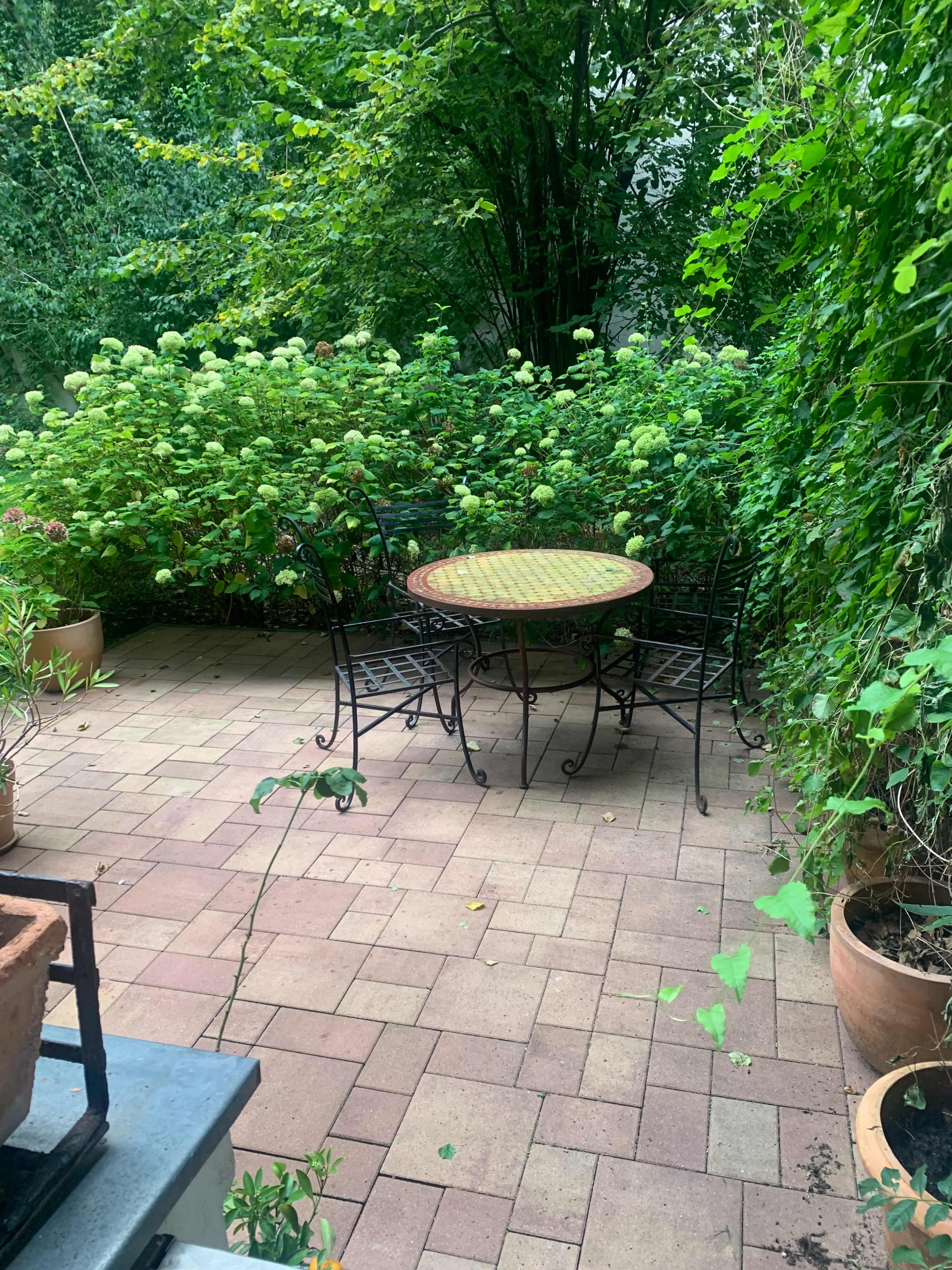 A wrought-iron table and chairs are positioned on a brick patio surrounded by lush green foliage and potted plants.