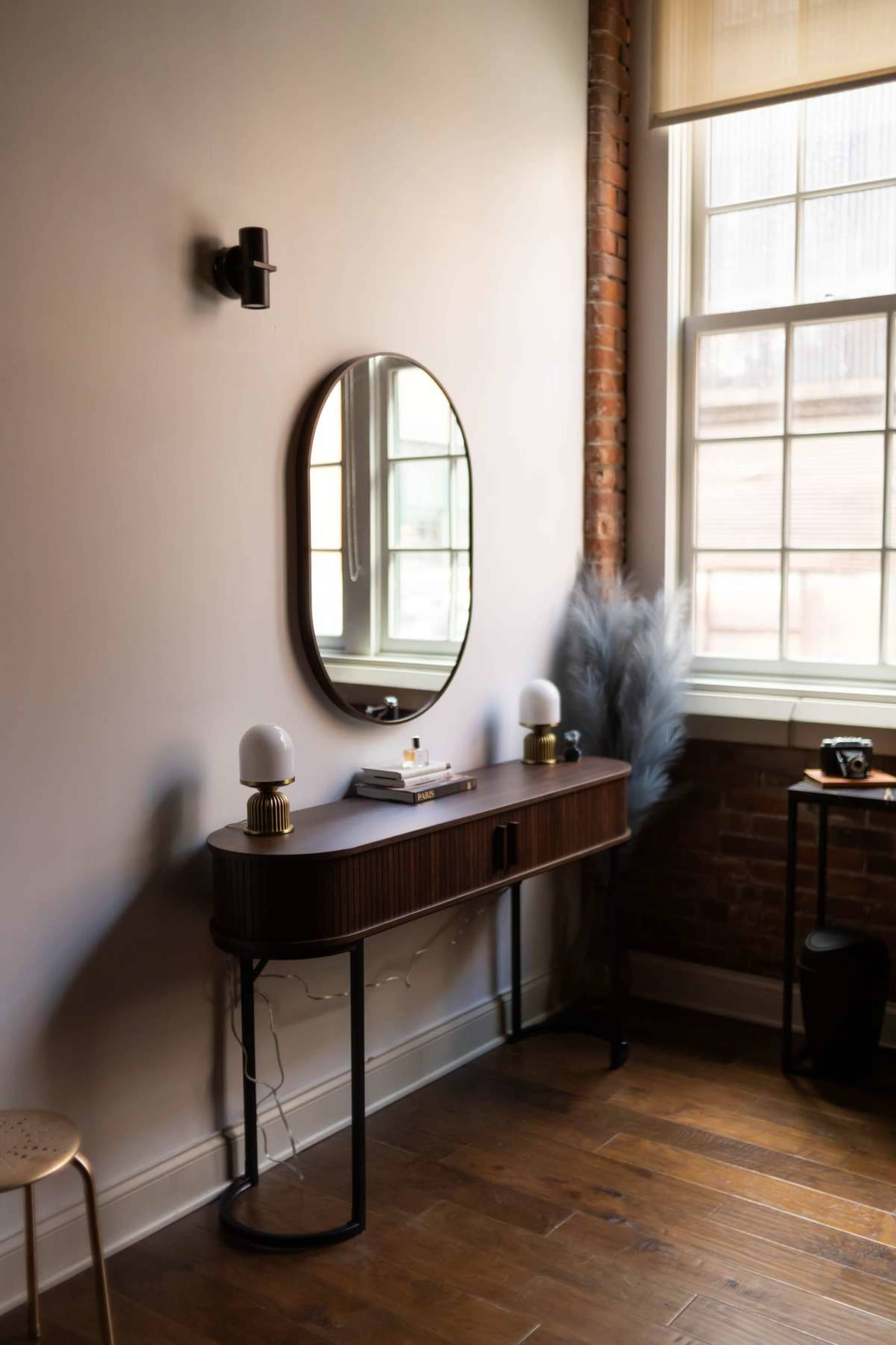 A wooden console table with lamps and decor sits against a wall with a large mirror reflecting the room's window.